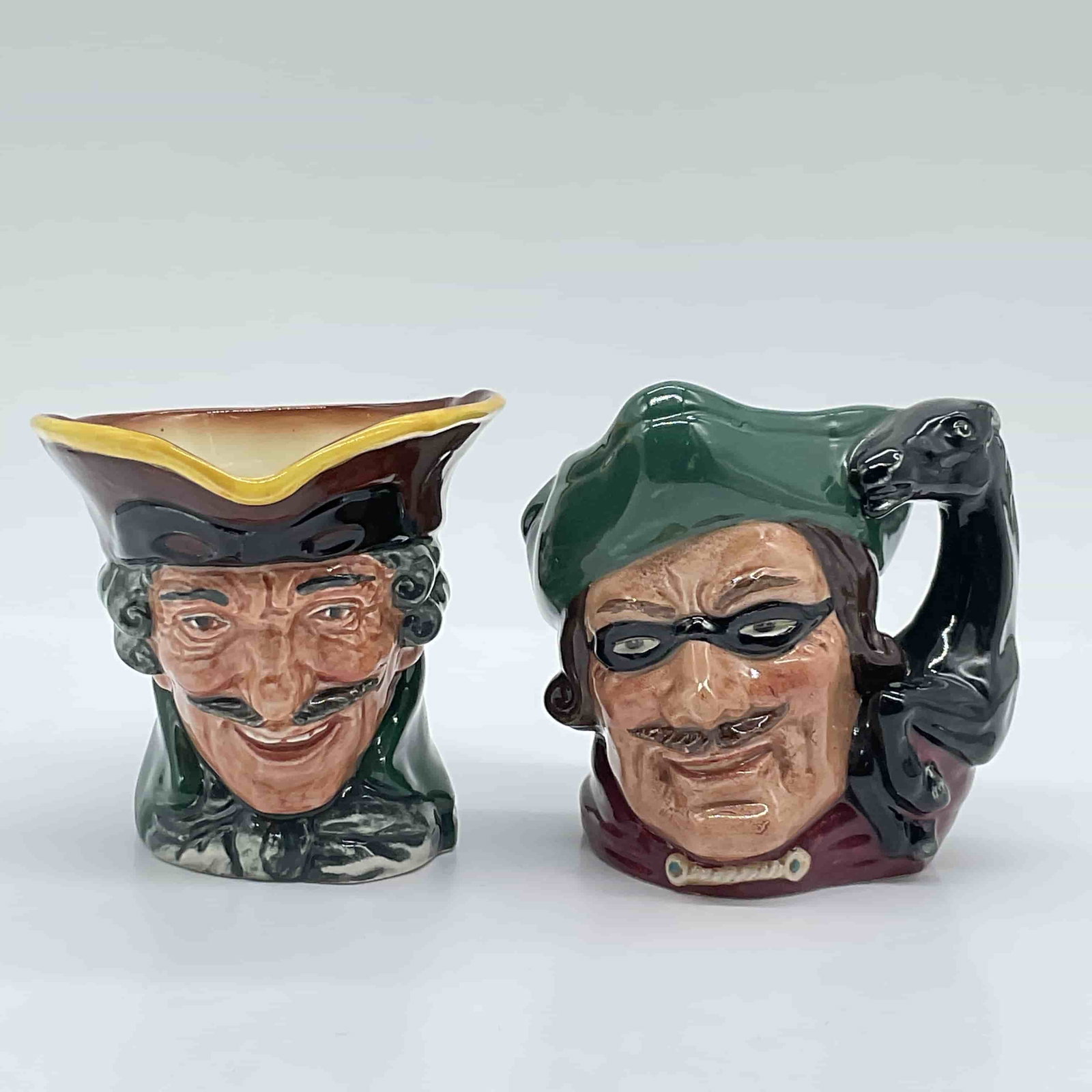 2pc Royal Doulton Character Jugs, Dick Turpin D6535 & D5618: Two Royal Doulton character jugs featuring the infamous English highwayman, Dick Turpin. This lot includes the D6535 jug, designed by David B. Biggs, with a tri-corner hat and a pistol-shaped handle,