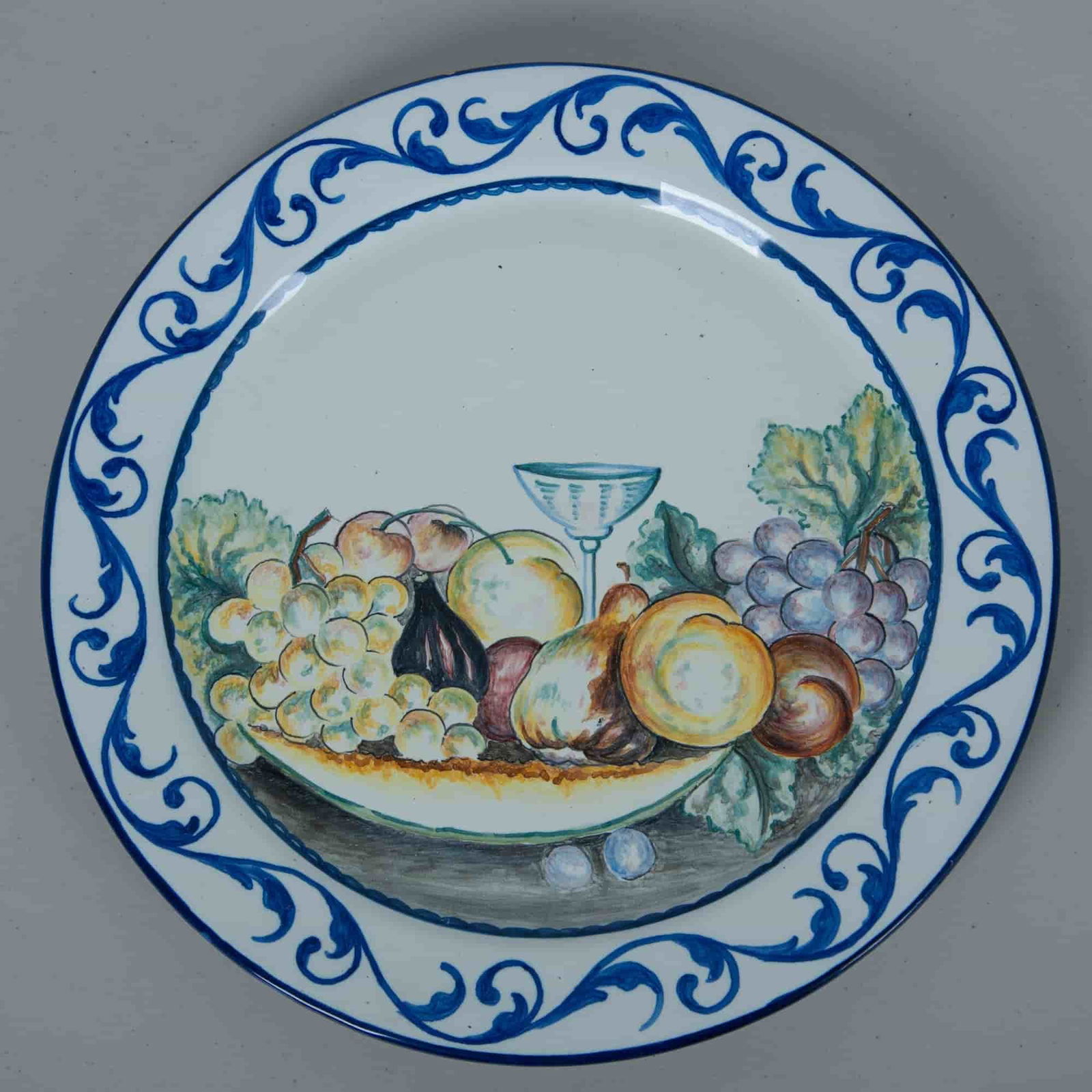 Vintage Large Ceramic Fruit Wall Plate: A white hand painted decorative plate depicting an array of fruits, leaves and a wine glass, with a dark blue scrollwork pattern around the rim. It has two holes on the back to allow for wall