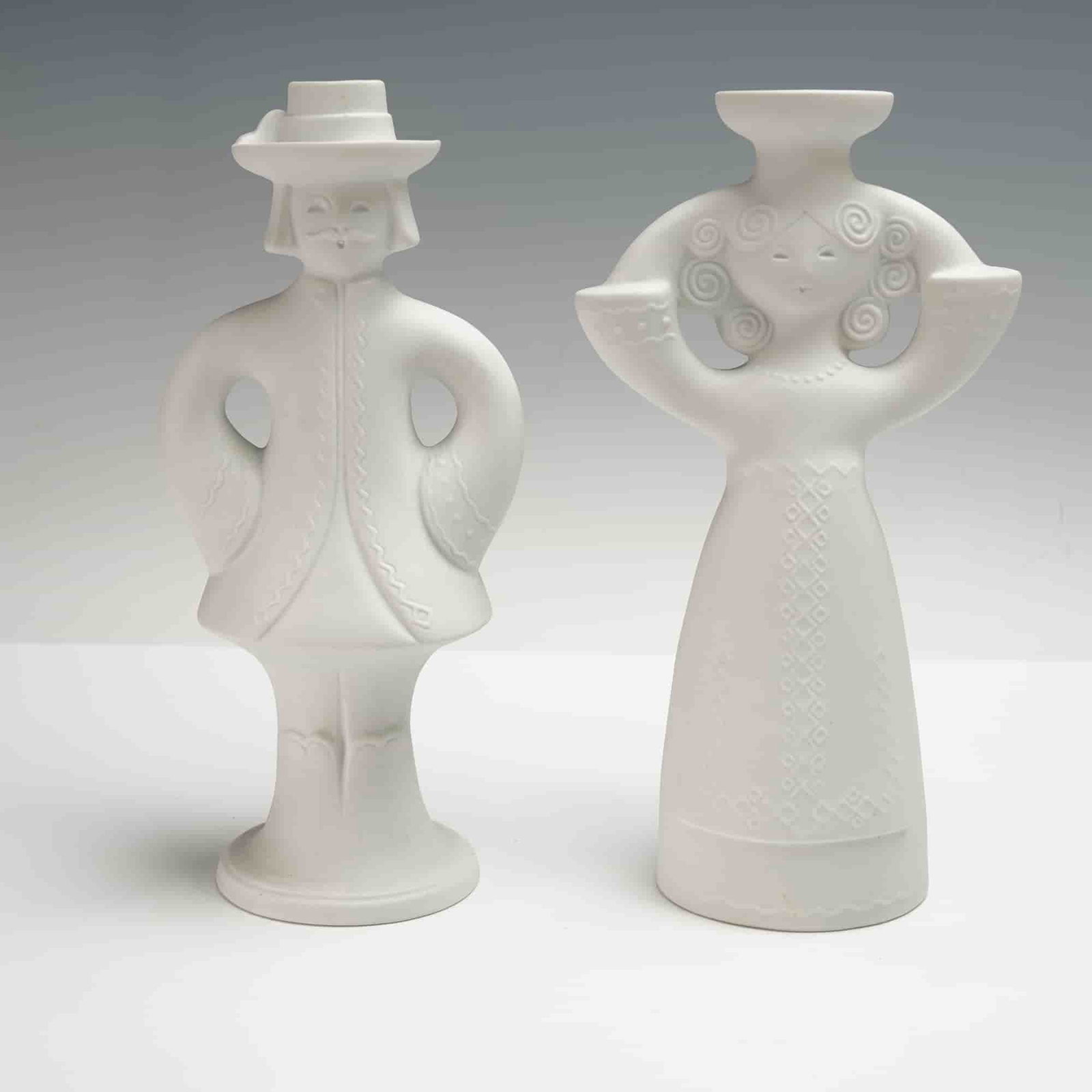 2pc AK Kaiser White Bisque Figural Candlestick Holders: Sculptural forms are characterized by flowing lines, pierced arm loops, and low-relief decorative elements, including spiral hair patterns and embossed costume details. The male figure wears a wide-br