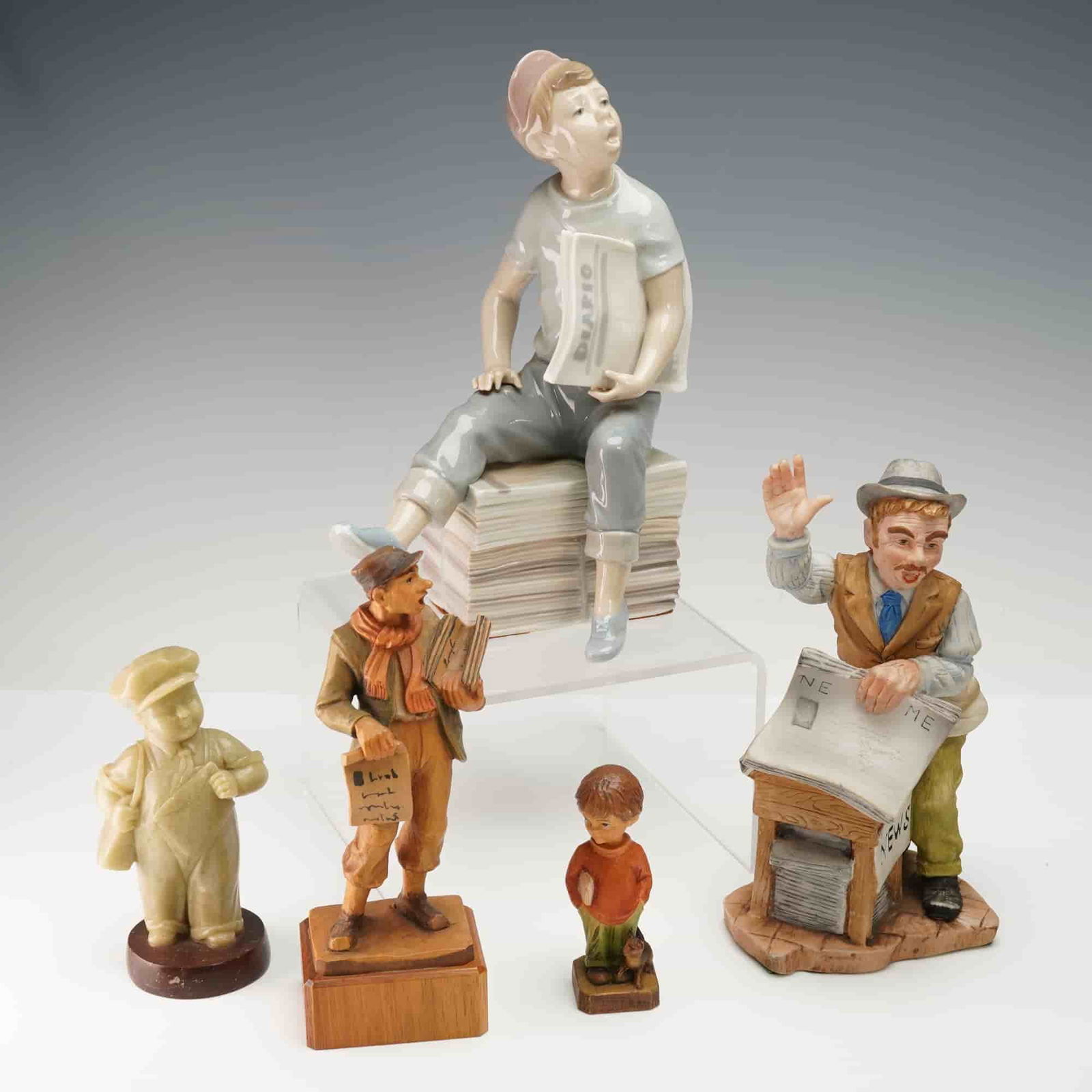 5pc Assortment of Newspaper Themed Figurines: Grouping of five newspaper-themed figurines in assorted materials including porcelain, ceramic, wood, and stone. Features a Nao by Lladro porcelain figure of a seated boy with newspapers and a