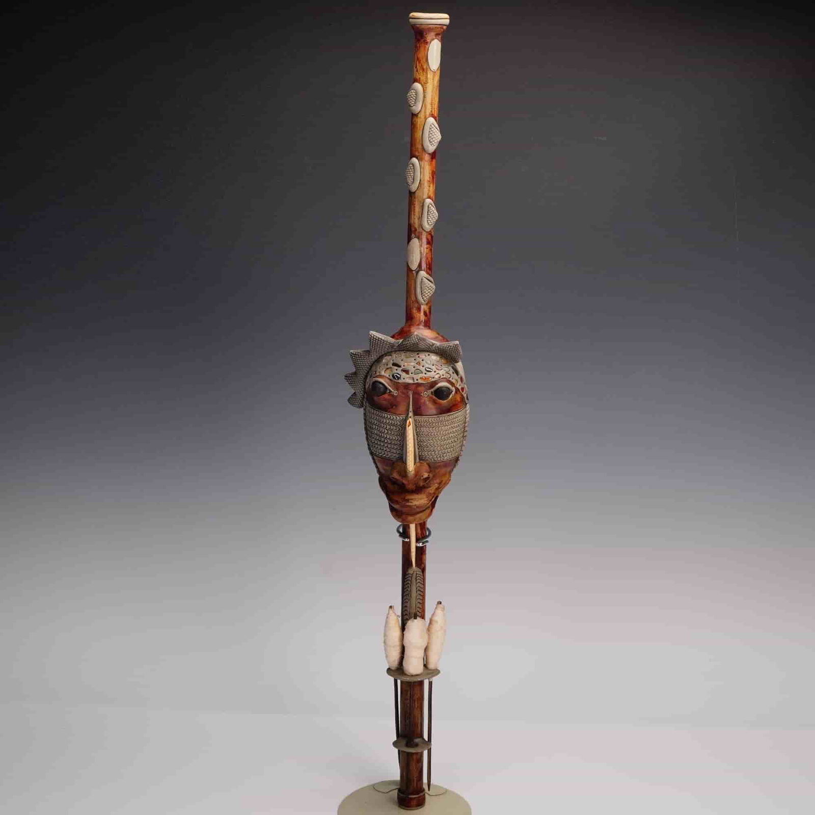 African Style Carved Wooden Staff Sculpture: Striking carved wooden staff sculpture featuring an elaborate mask face with intricate patterns and raised elements, accented by inset shells and beads, creating expressive eyes and a prominent