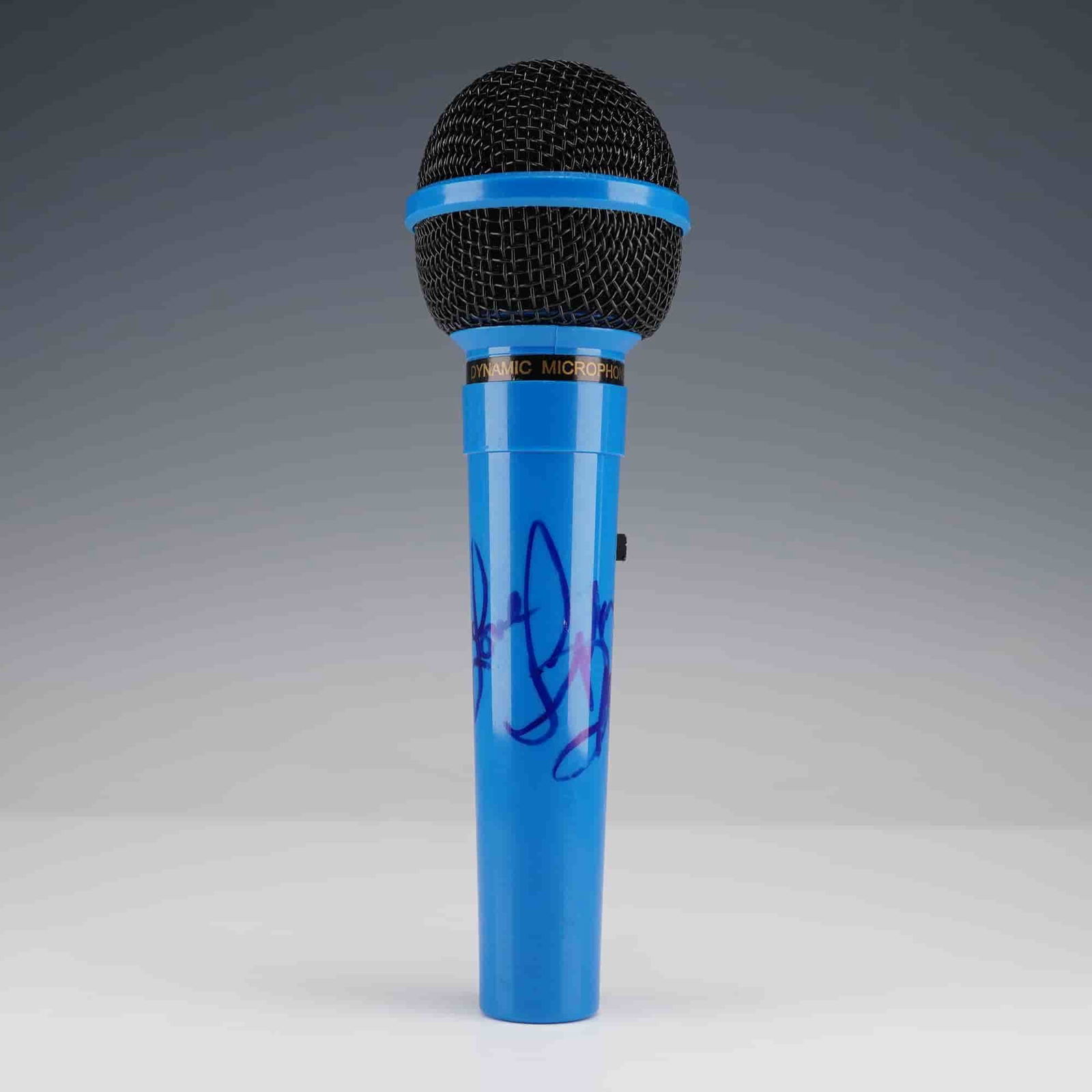 Autographed Blue Microphone: Bright blue SPM-03 dynamic microphone with black grille, featuring a large autograph in blue marker across the handle. The signature is stylized and not fully legible. Fitted with on/off switch and