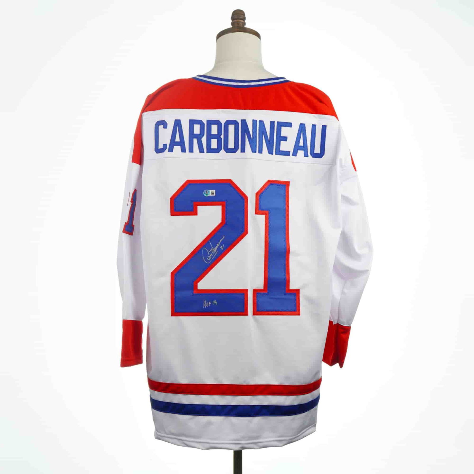 Guy Carbonneau Signed Montreal Canadiens Style Custom Hockey Jersey HOF 19: A single signed jersey styled after a Montreal Canadiens uniform, constructed of white synthetic fabric with red shoulder yokes and red and blue striping on the cuffs, hem, and V-neck collar. The