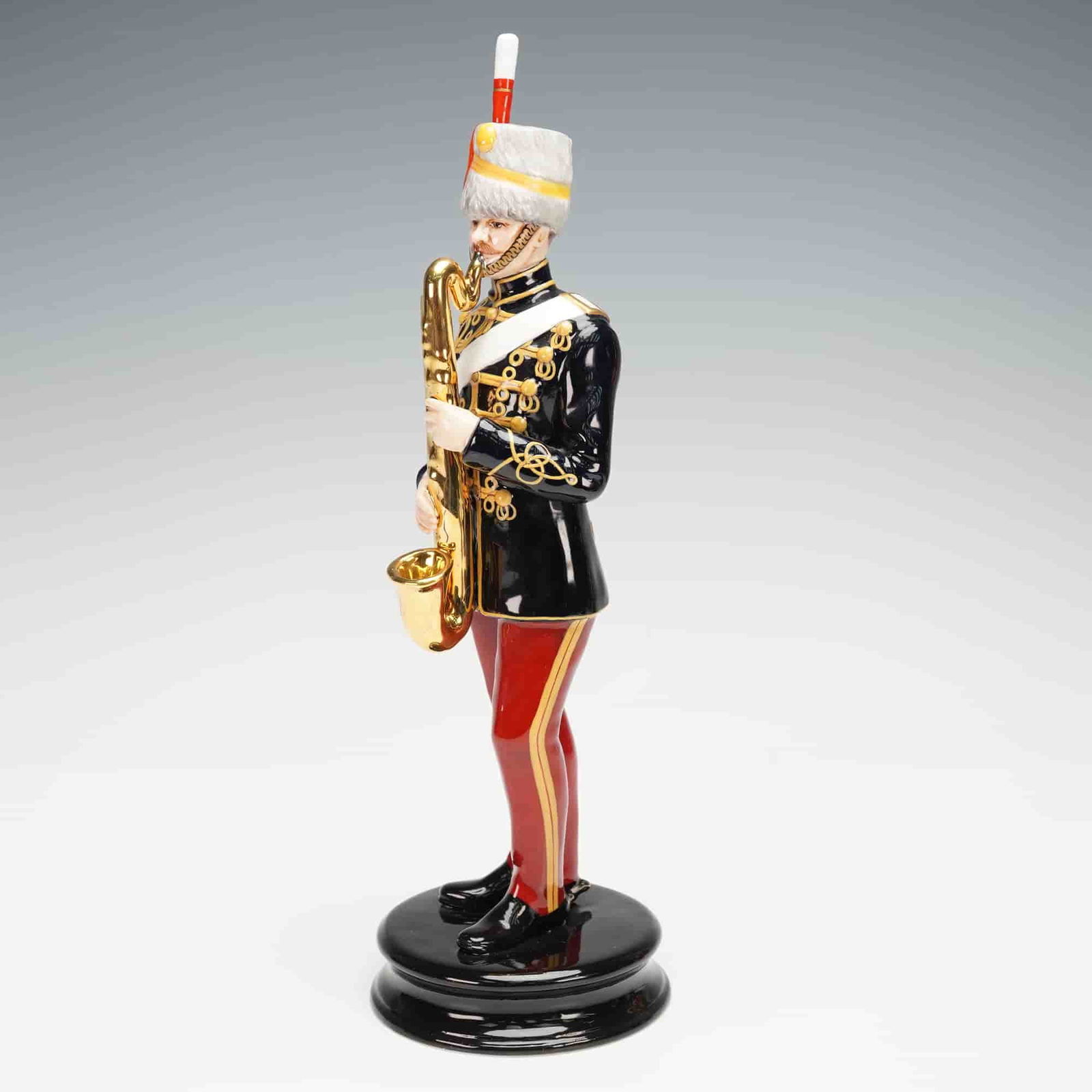 Michael Sutty Porcelain Sculpture, 11th Hussars: A hand painted porcelain figure showing a member of the 11th Hussars in a dark coat with gold trim, red trousers, and a tall white busby, holding a long saxophone in front of him as he plays it while