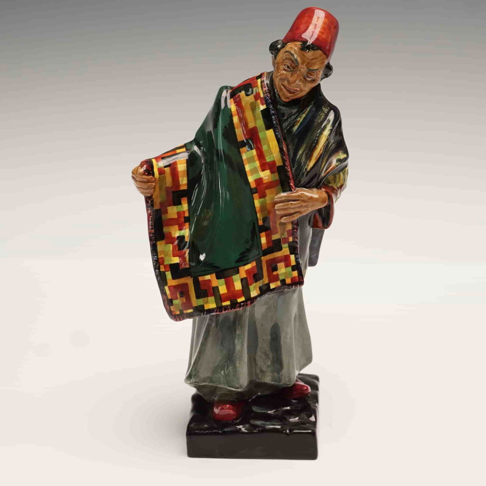 Royal Doulton Porcelain Figurine, Carpet Seller HN1464: This striking Royal Doulton figurine titled Carpet Seller (HN1464) captures the vibrant essence of a traditional market merchant. Dressed in flowing robes and a bright red fez, the figure is portrayed
