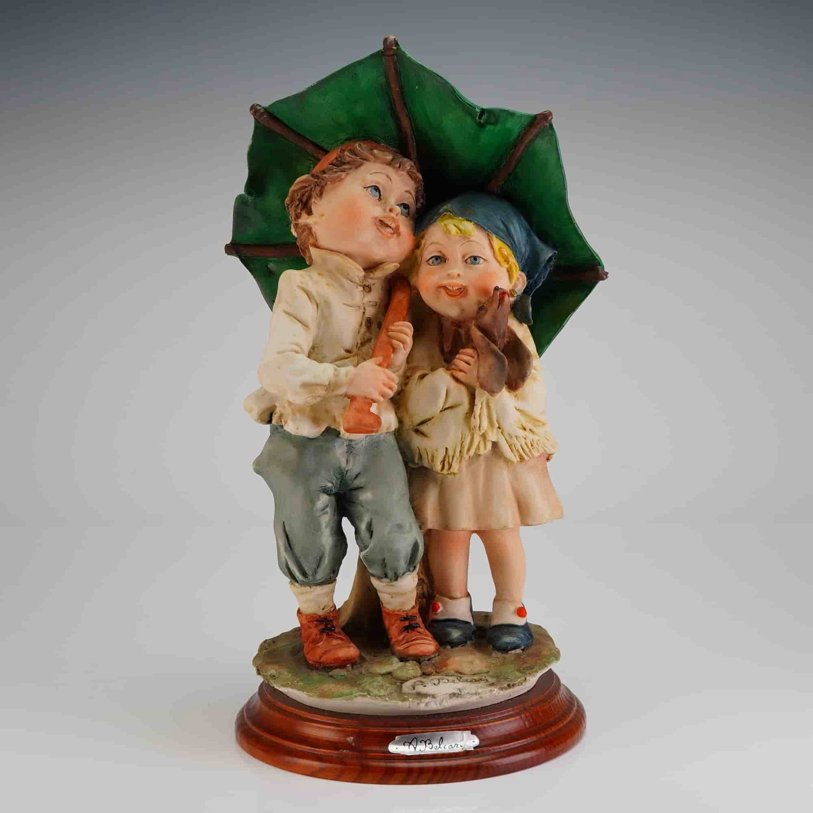Capodimonte A. Belcari Figurine Children Under Umbrella: A sentimental genre Capodimonte figurine signed by the artist A. Belcari. The sculpture, executed in cold-painted composite material, depicts two cherubic children, a boy and a girl, standing close to