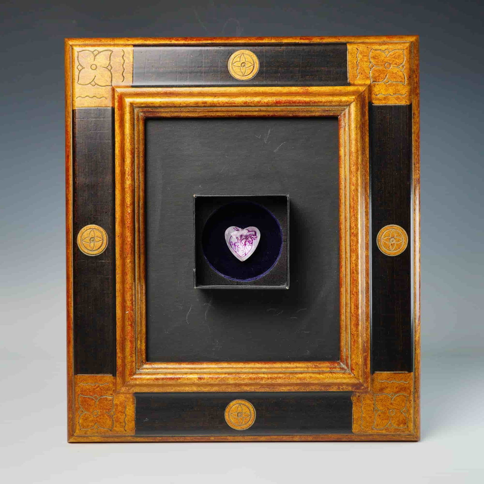 Framed Heart Medallion Art Work: A single Framed Art work featuring a heart medallion. The object is presented in a custom wooden frame detailed with speckled gold-tone borders and contrasting ebonized central bands. The frame is
