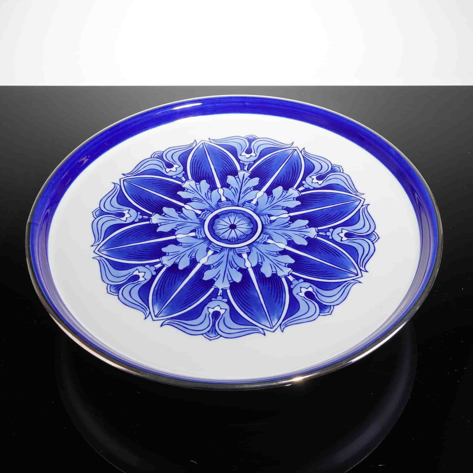 Bombay Blue and White Porcelain Pedestal Cake Stand: The stand is executed in glazed white ceramic and features a modern decorative style. The top surface is decorated with a complex, symmetric eight-lobed floral medallion rendered in blue transfer-prin