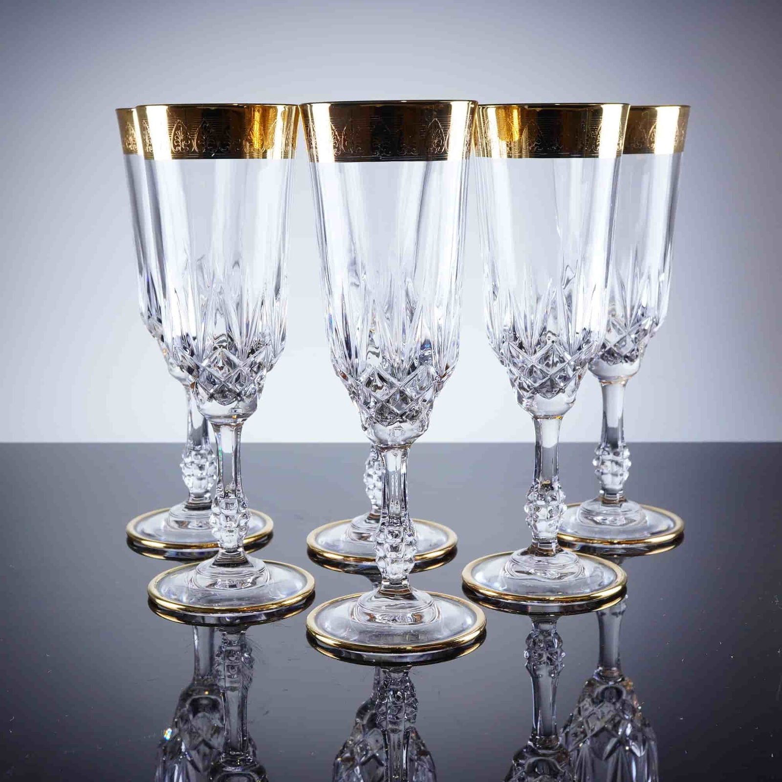 6pc Gold Rim Crystal Champagne Flutes: A collection of six clear crystal champagne flutes, featuring a tall, slender bowl, extensively decorated with deep diamond-point cutting. The upper rim of the bowl is finished with a wide band of app