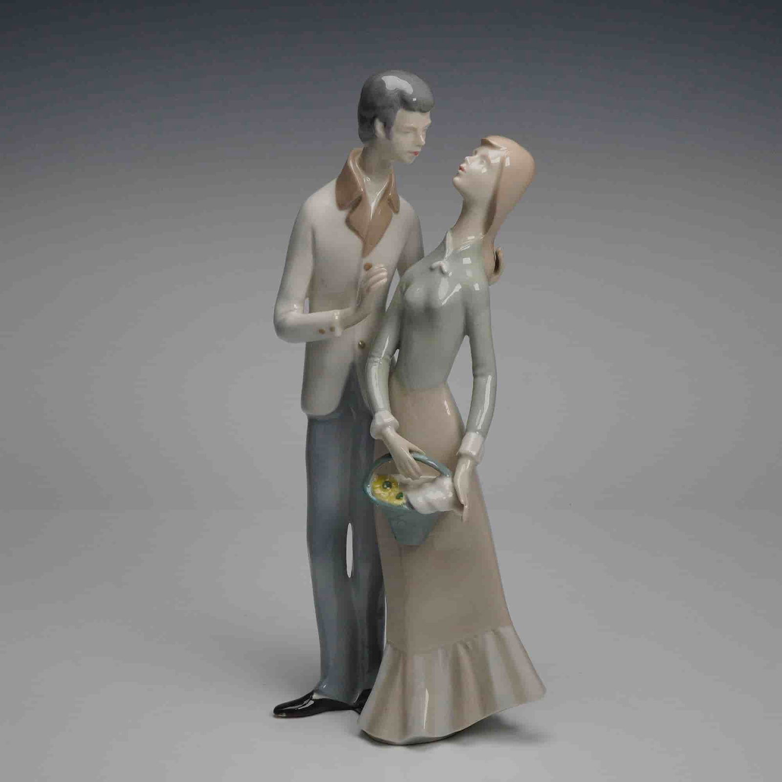 GDR Lippelsdorf Young Couple Porcelain Figurine: A glazed porcelain figurine from Lippelsdorf in the German Democratic Republic depicting a young couple standing closely together, the woman holding a small basket of yellow flowers and dressed in a