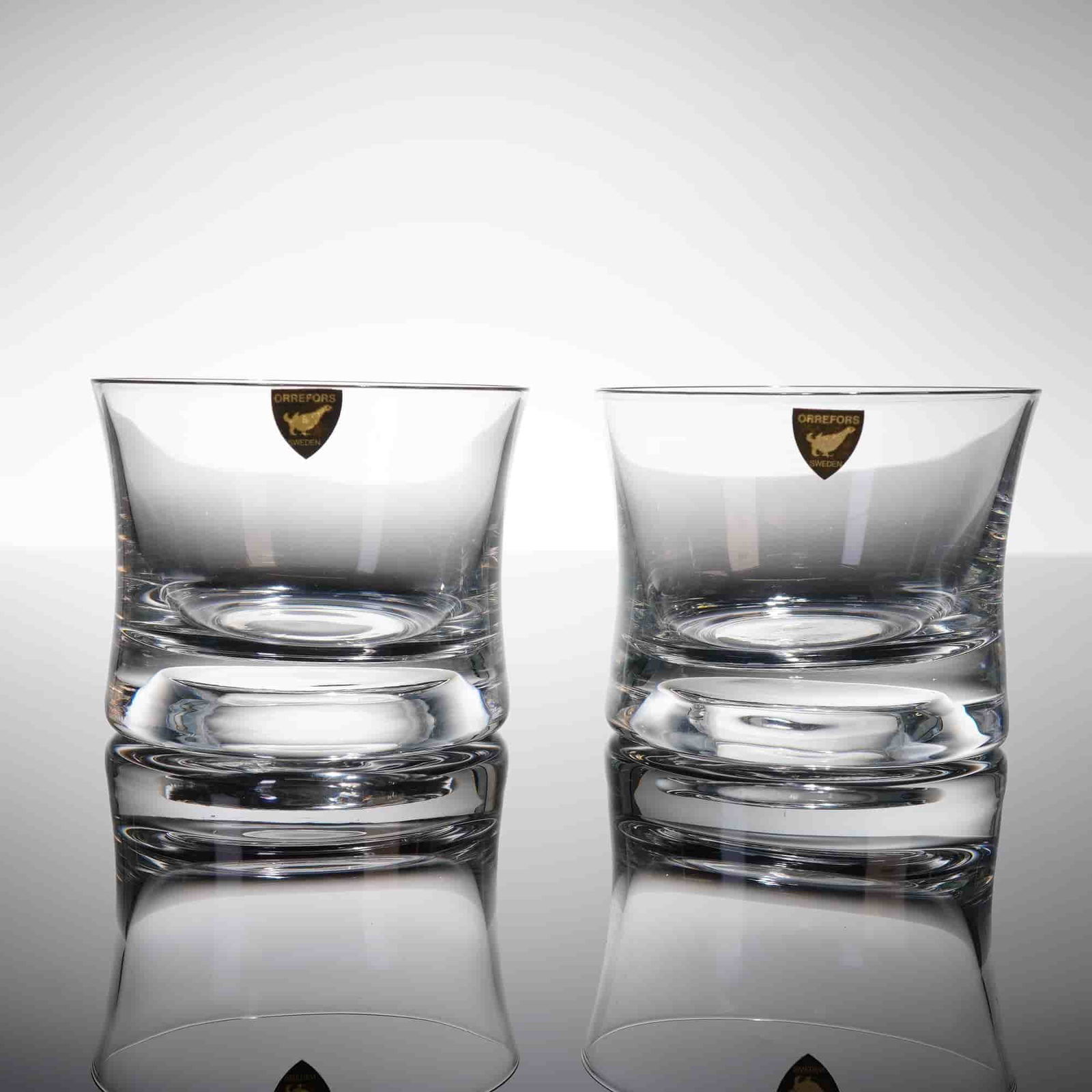 A Pair of Orrefors Clear Crystal Rocks Glasses: A pair of heavy clear crystal tumblers, commonly used as rocks glasses. The glasses are characterized by a thick, solid base and gently curving walls that iare slightly outward toward the rim. Both