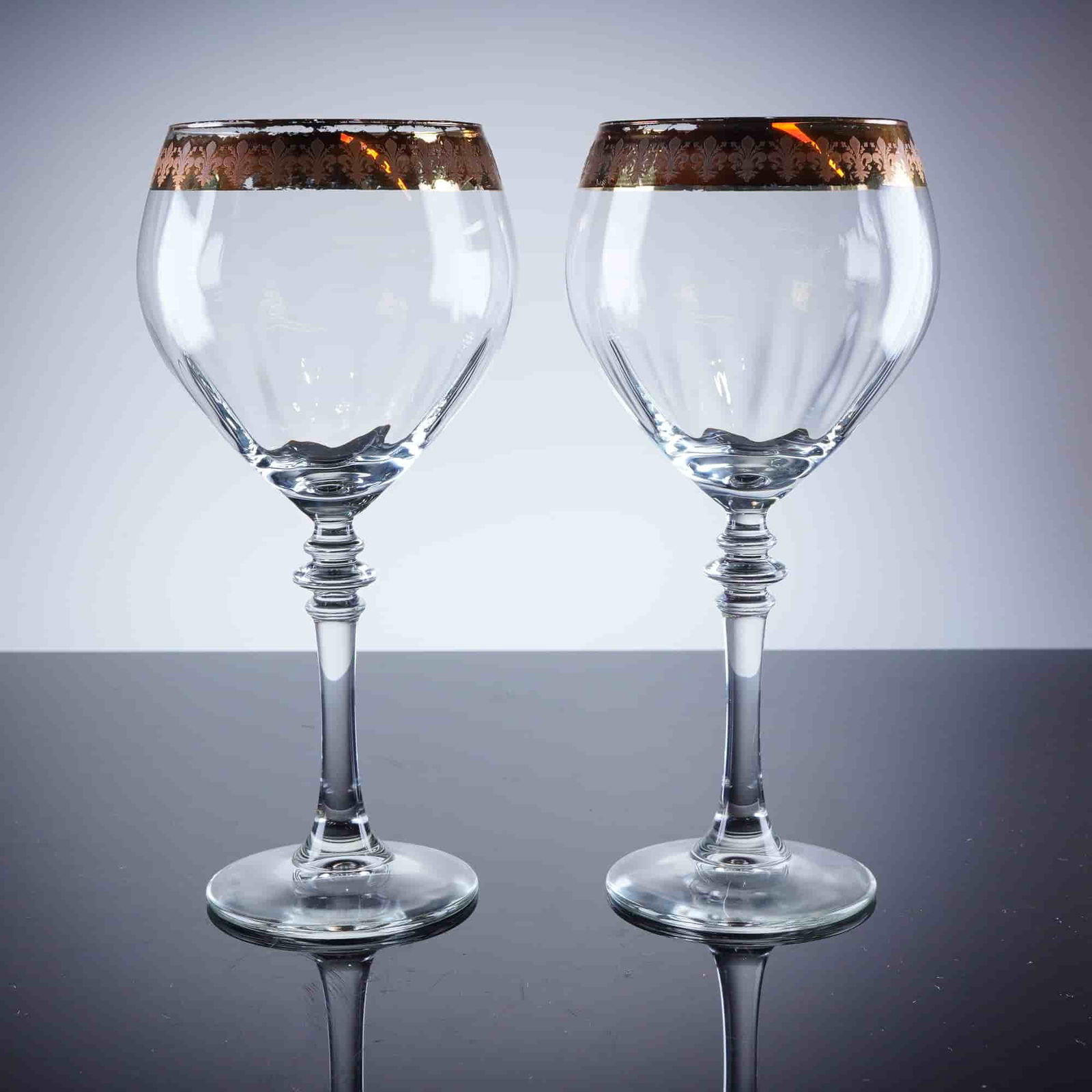 2pc Gold Rim Fleur De Lis Wine Glasses: A pair of stemmed clear crystal wine glasses featuring a classical revival style with tall, knopped stems and slightly tapered, tulip-shaped bowls. The bowls are detailed with vertical facet cutting