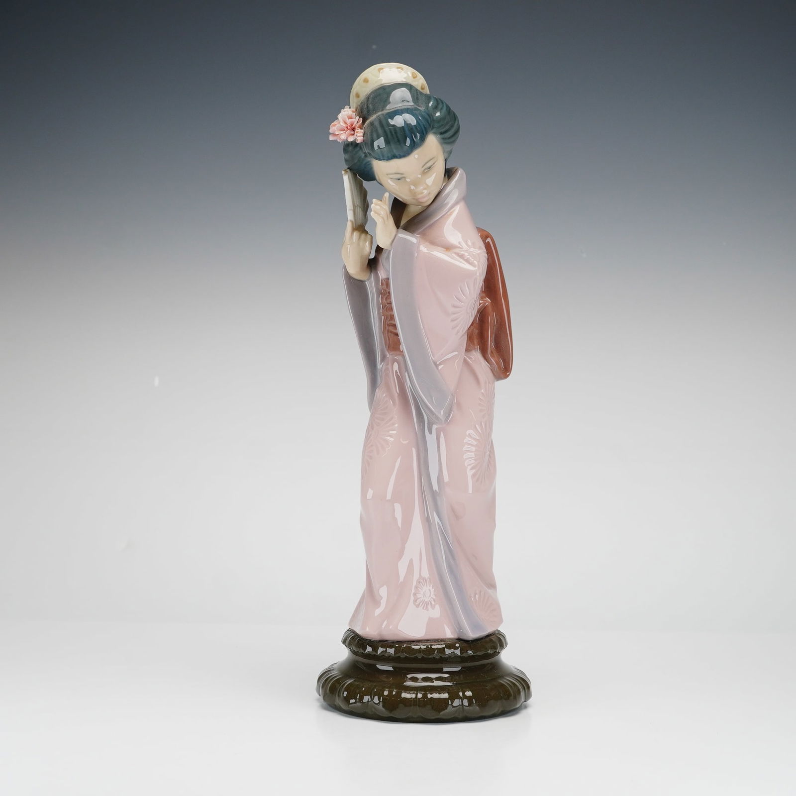 Lladro Porcelain Figurine, Chrysanthemum 1004990: This fine glazed porcelain figurine depicts a standing Japanese woman in a traditional pale pink kimono with a subtle floral pattern, lavender-gray trim, and a brown obi. She is styled with a traditio