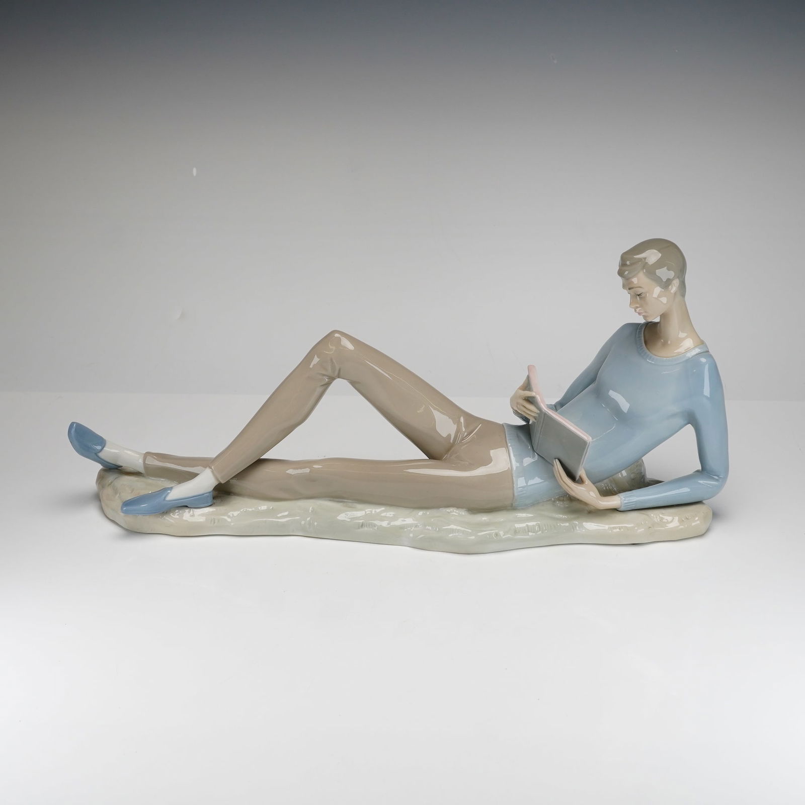 Lladro Porcelain Figurine, Boy with Book 1001024: This is a large, hand-painted glazed porcelain figurine depicting a young man in a relaxed, reclining pose upon a rocky-textured base. He is depicted reading an open book, dressed in a pale blue sweat