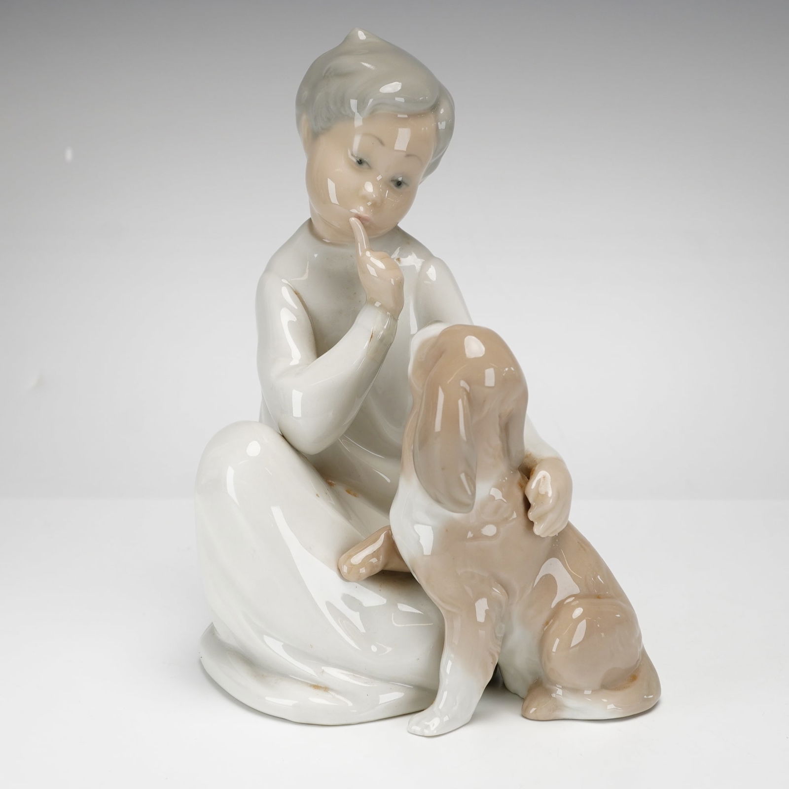 Lladro Porcelain Figurine, Boy with Dog 1004522: This glazed porcelain figurine features a young boy in a white nightshirt seated on the floor, with his finger to his lips while a small spaniel-type puppy leans against him. It is finished in the cla