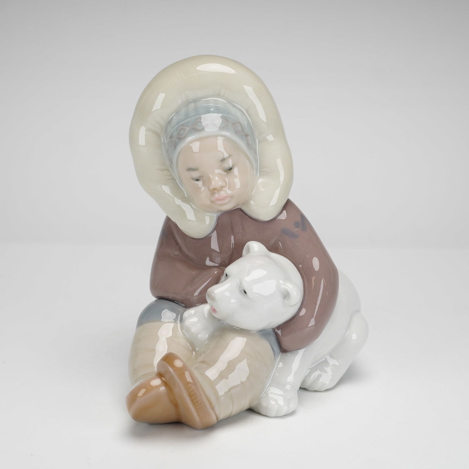 Lladro Porcelain Figurine, Eskimo 1001195: This is a glazed porcelain figurine depicts a young boy wearing a traditional hooded parka and patterned cap, sitting and affectionately embracing a white polar bear cub. The figurine is finished in a