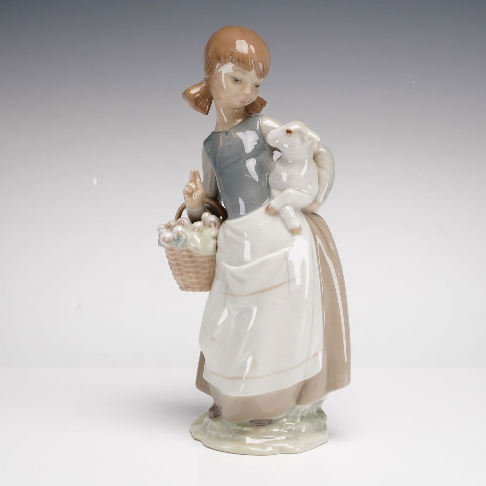 Lladro Porcelain Figurine, Girl with Lamb 1004835: A polychrome glazed porcelain figurine depicting a young shepherdess with pigtails in traditional rustic attire, featuring a gray bodice, white apron, and tan-colored skirt. She holds a small lamb in
