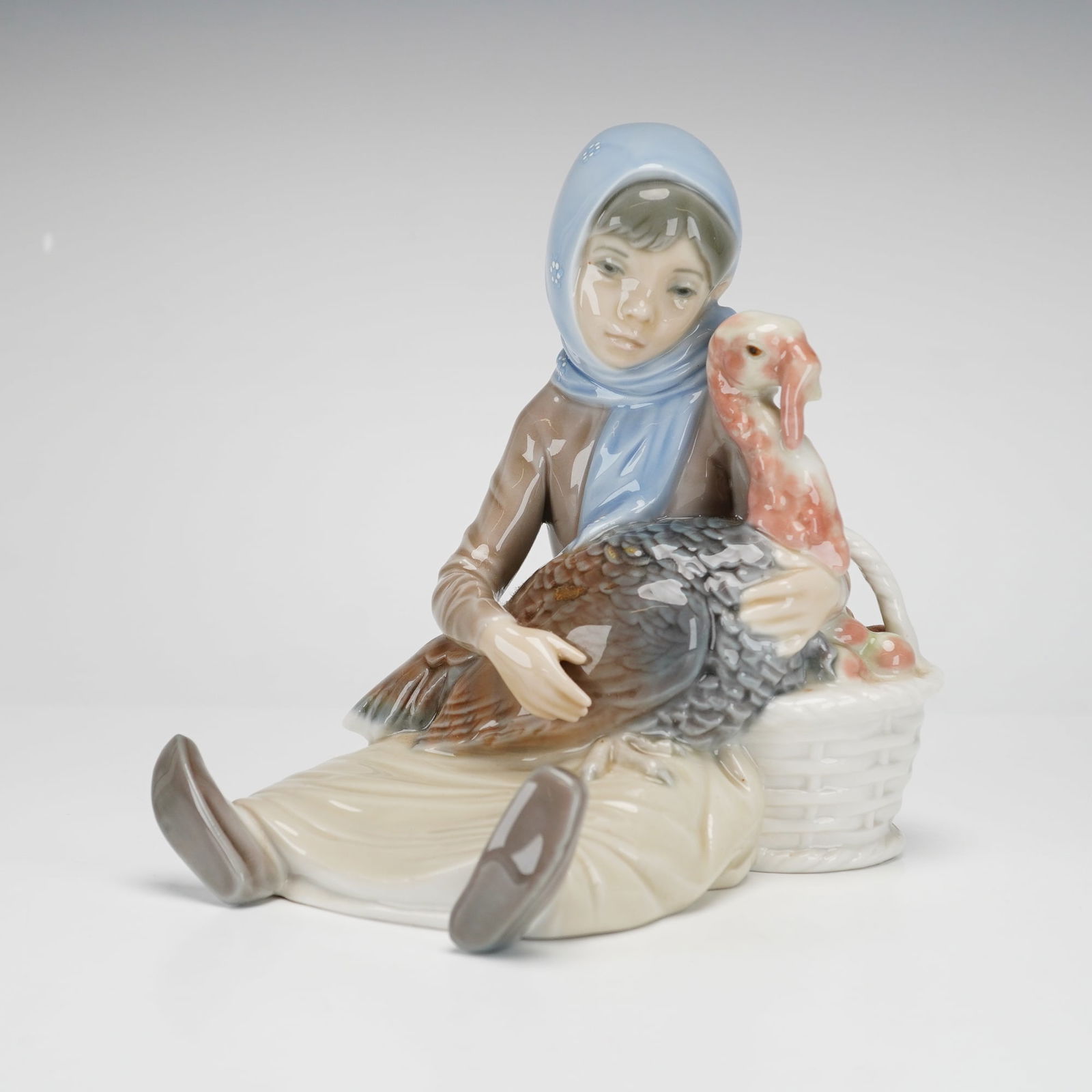 Lladro Porcelain Figurine, Girl with Turkey 1004569: This is a glazed polychrome porcelain figurine depicting a young girl wearing a pale blue headscarf and brown jacket seated while holding a large turkey with detailed plumage in shades of brown, gray,