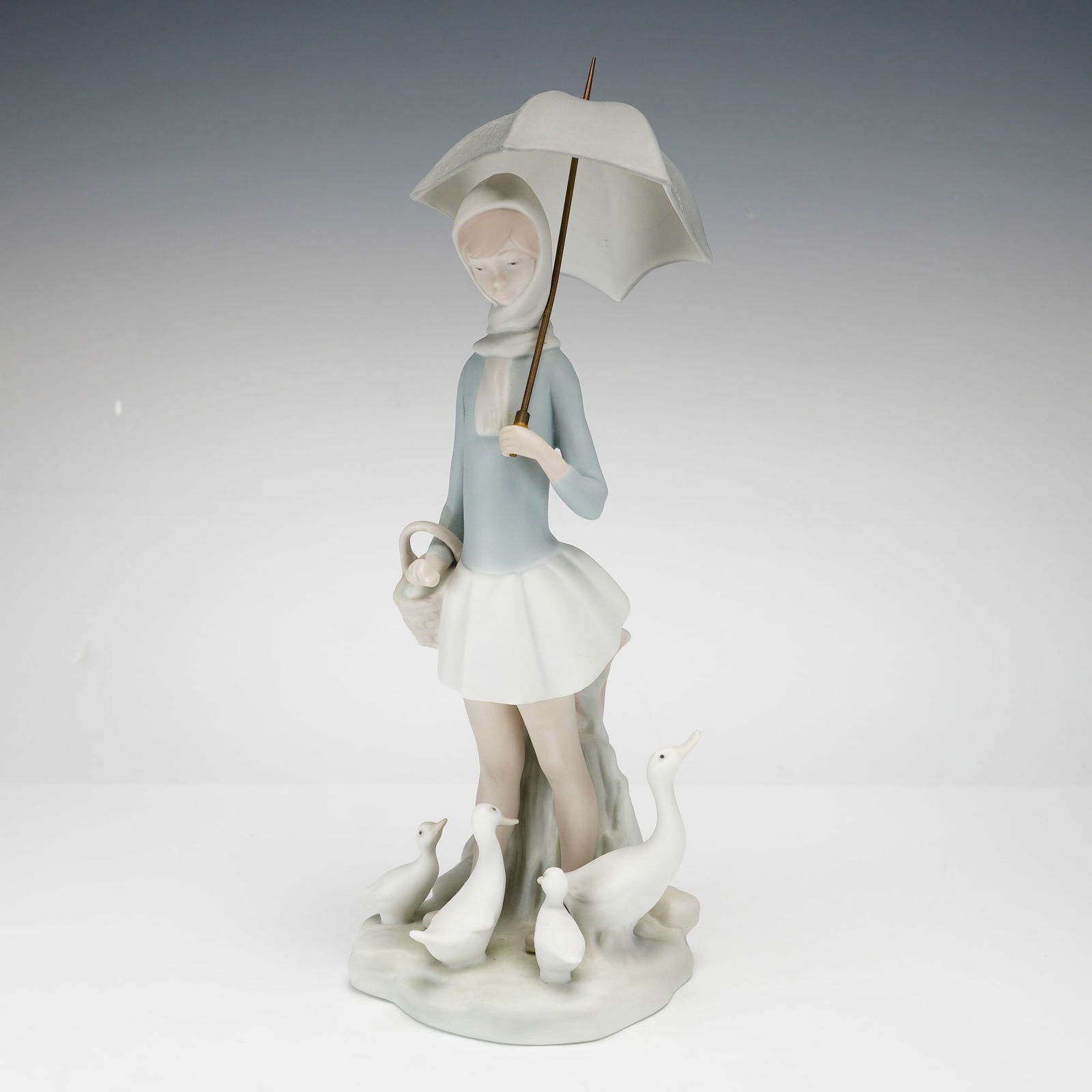 Lladro Porcelain Figurine, Girl with Umbrella and Geese 1014510: This Lladro matte porcelain figurine depicts a young girl in a soft blue hooded jacket, white scarf, and pleated skirt, holding a wicker basket and a gray umbrella with a metal shaft. Four white geese