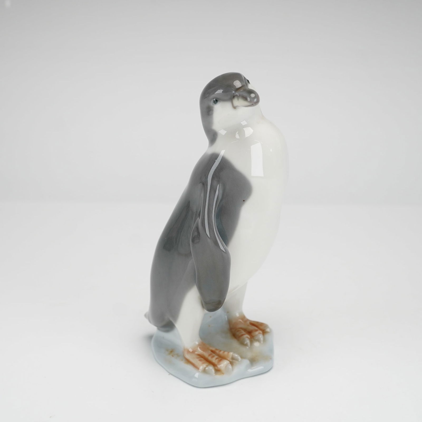 Lladro Porcelain Figurine, Penguin 1005249: This is a glazed porcelain figurine produced by the Spanish factory Lladro, depicting a standing penguin in a naturalistic pose with its head tilted slightly upward and to the right, perched upon a sm