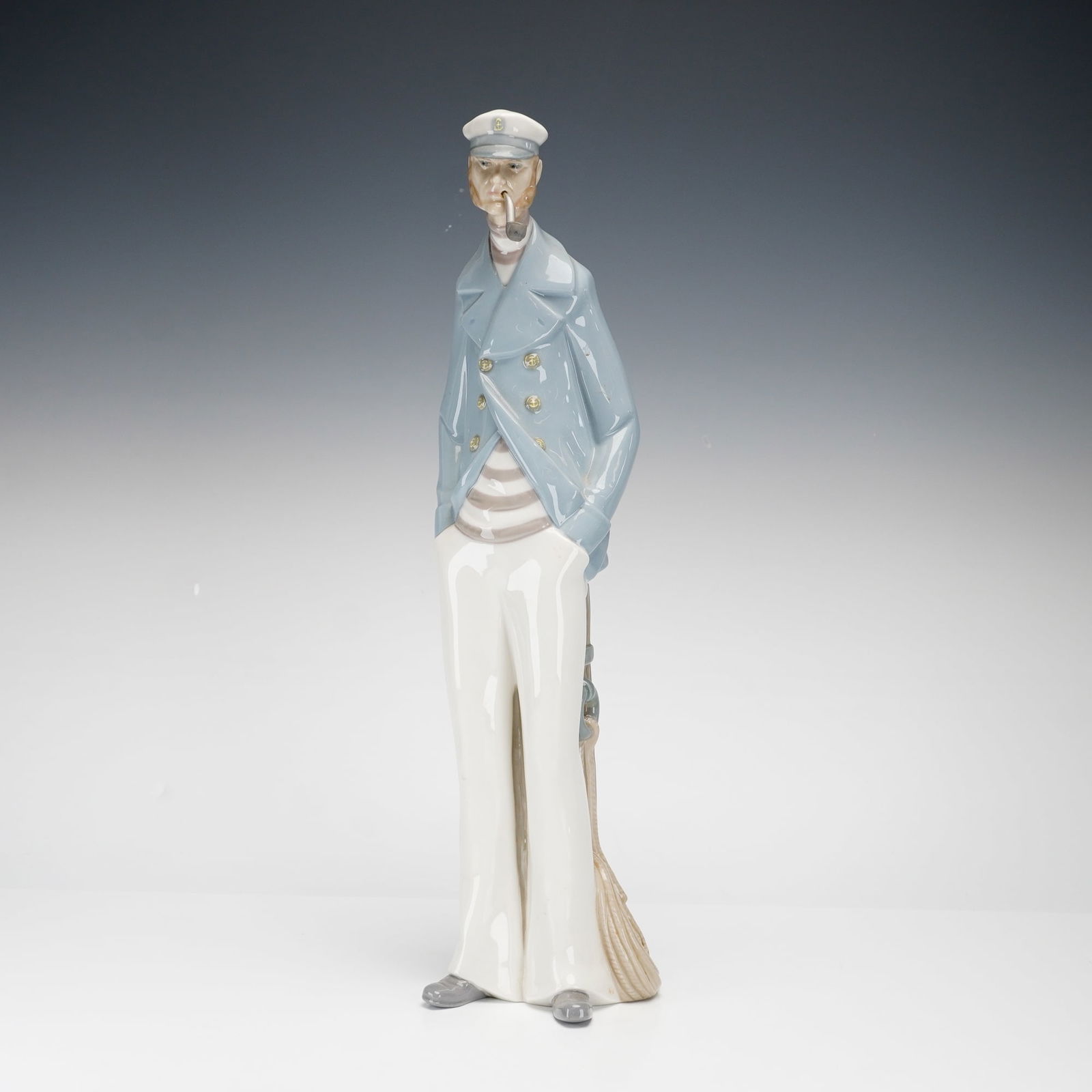 Lladro Porcelain Figurine, Sea Captain 1004621: This is a tall, slender glazed porcelain figurine depicting a bearded maritime officer smoking a pipe and leaning casually against a wooden mooring piling with a coiled rope at the base. The officer i