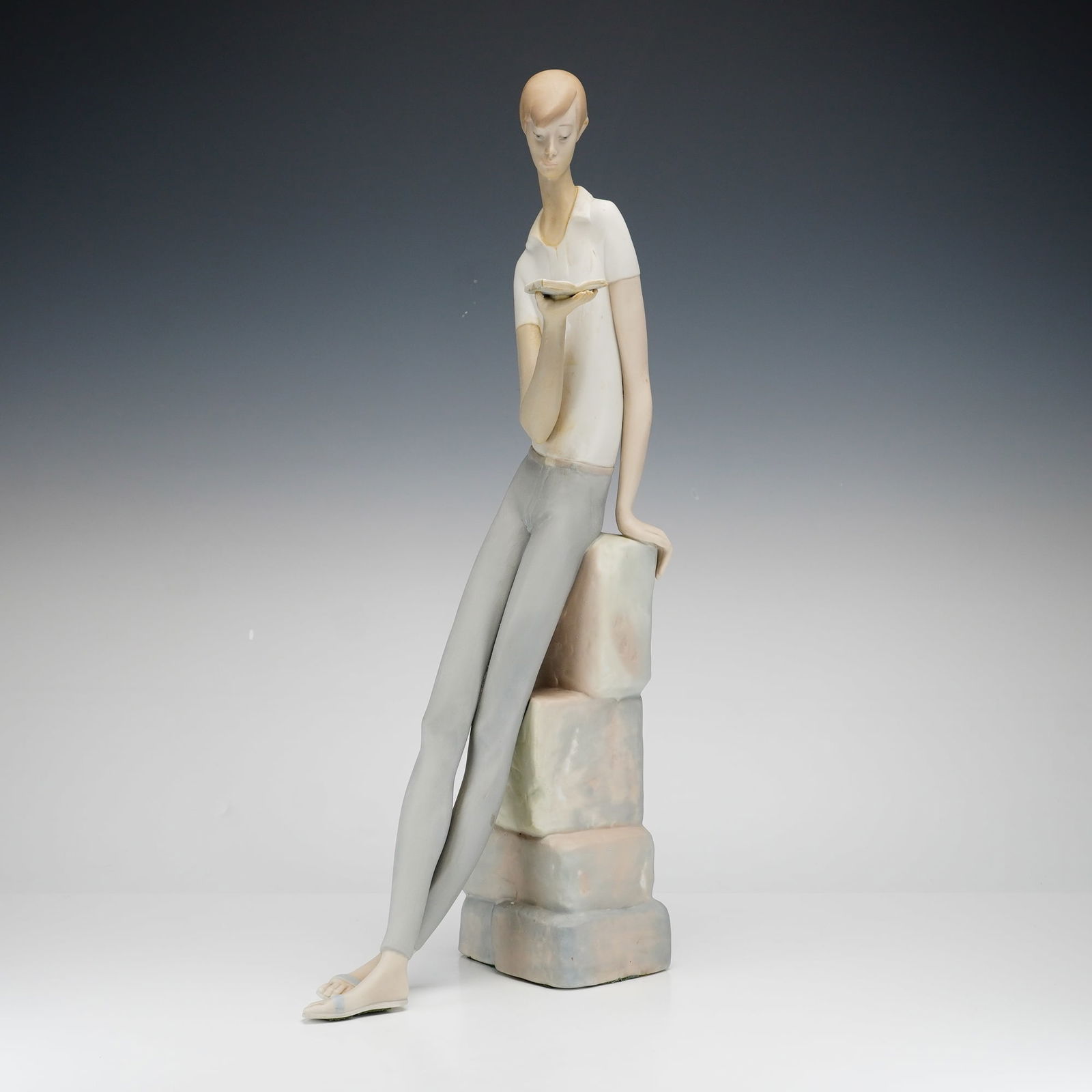 Lladro Porcelain Sculpture, Boy Student 1014517: A Lladro porcelain sculpture depicting a stylized, elongated young man in contemporary attire leaning against a stack of square blocks while looking down at a book. The sculpture features a matte fini