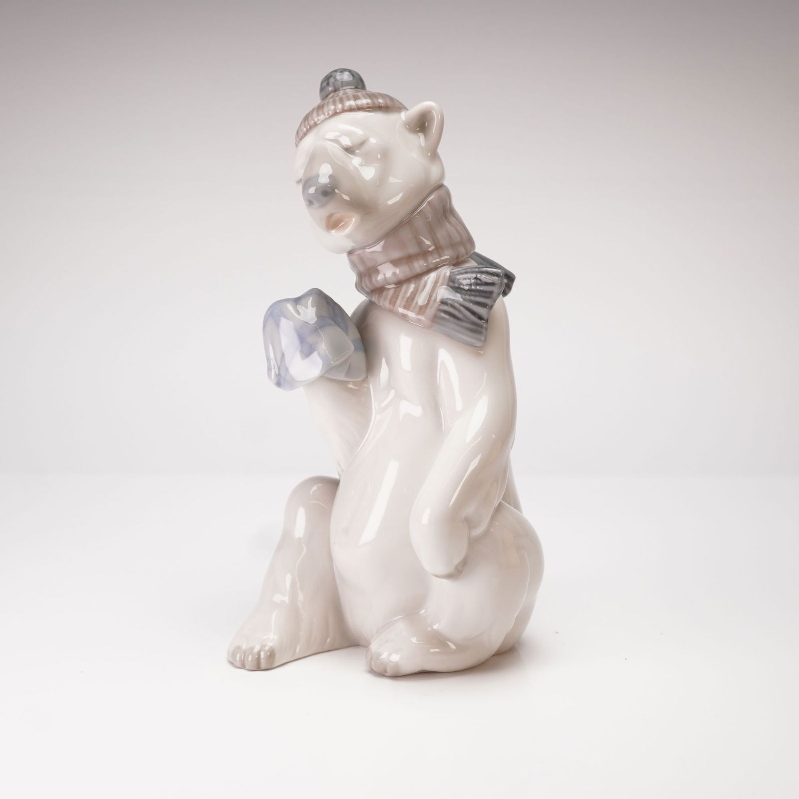 Lladro, Painful Bear Figurine 1005021: This whimsical Lladro fine glazed porcelain figurine, titled Painful Bear model 1005021, depicts an anthropomorphized seated polar bear. The bear is shown wearing a mauve and white striped knit beanie