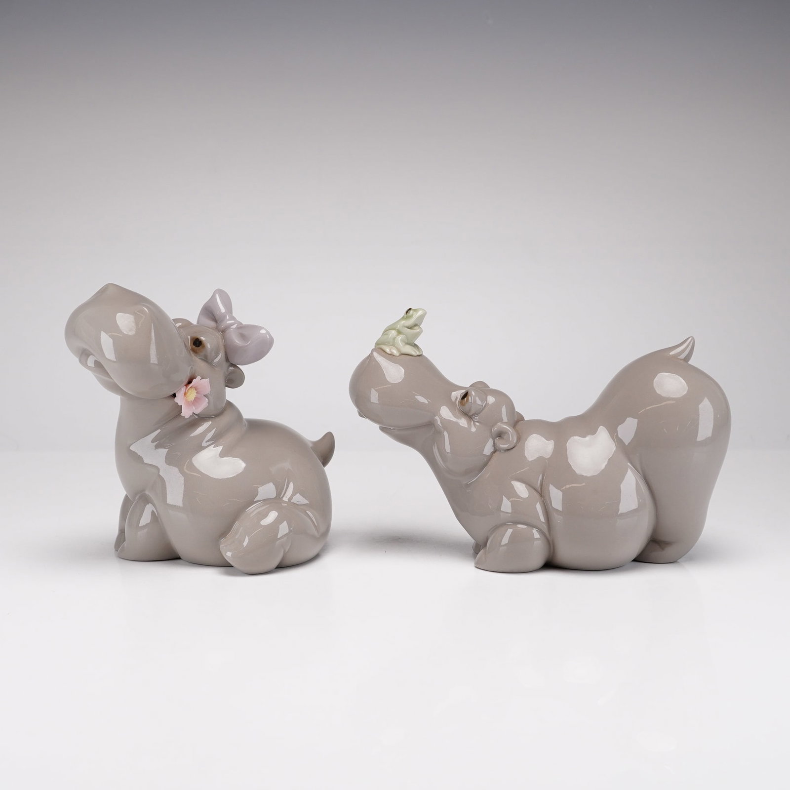 2pc Nao by Lladro Porcelain Figurines, Hippo in Love and Hippo with Frog: This lot features two glossy glazed porcelain figurines manufactured by Nao. The first figurine, model number 1321, titled Hippo in Love, depicts a seated hippopotamus wearing a lavender bow and holdi