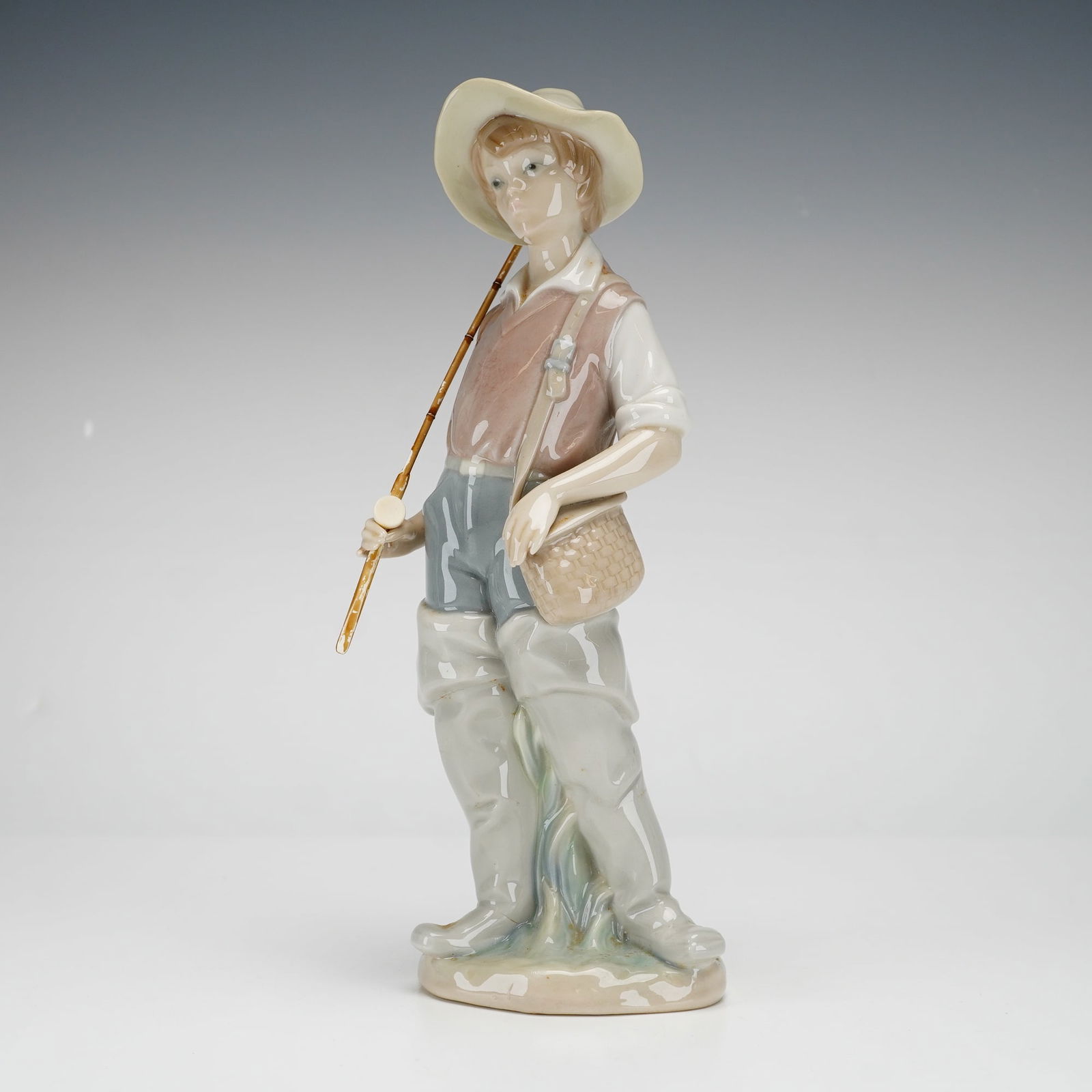 Lladro Porcelain Figurine, Going Fishing 1004809: This Lladro glazed porcelain figurine, titled Going Fishing, was designed by sculptor Salvador Furio. The piece depicts a young boy dressed for fishing in a wide-brimmed sun hat, a mauve vest over a w