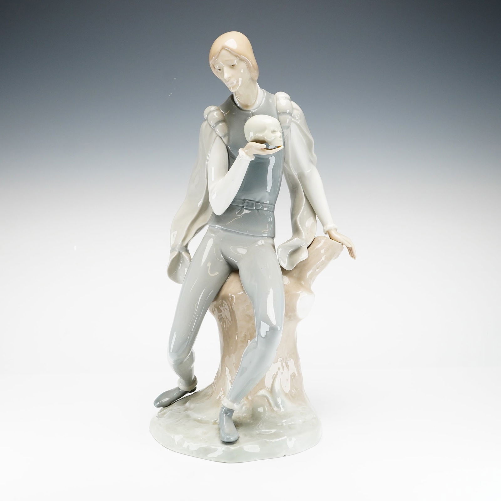 Lladro Porcelain Sculpture, Hamlet 1004729: This glazed porcelain sculpture by Lladro depicts the Shakespearean character Hamlet in a pensive, seated pose. Designed by sculptor Alfredo Ruiz, the figure is shown leaning against a stylized tree s