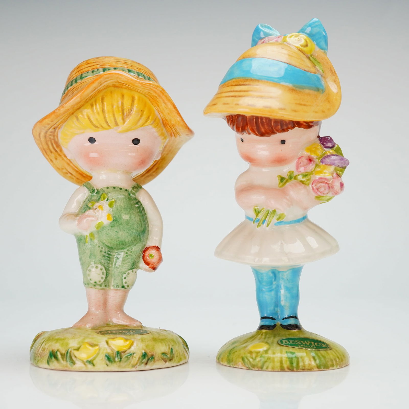 2pc Beswick Joan Walsh Anglund Ceramic Figurines: A pair of glazed ceramic figurines produced by Beswick, featuring the illustrative style of artist Joan Walsh Anglund. The collection includes a figure of a boy wearing green overalls and a broad-brim