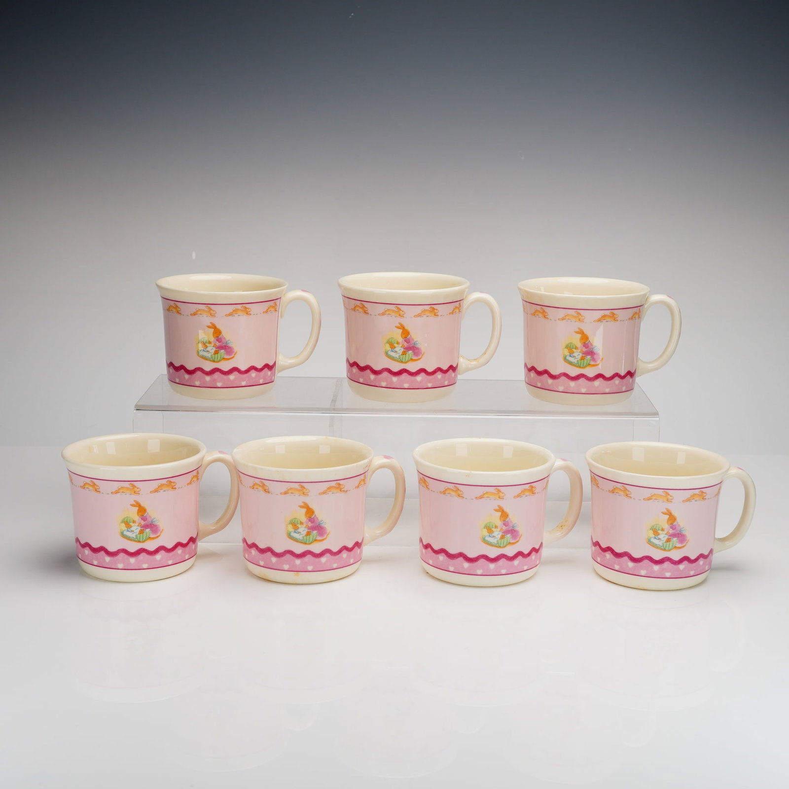 7pc Royal Doulton Bunnykins Sweet Hearts Hug A Mugs Set: A collection of seven Royal Doulton ceramic mugs in the Bunnykins Sweet Hearts pattern. Each mug features a cream-colored body with a pink wrap-around band decorated with a white heart pattern and cen