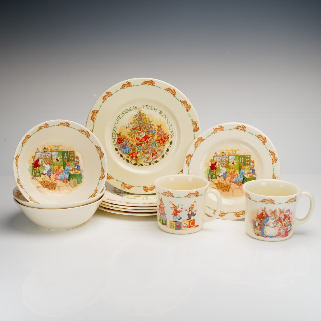 11pc Royal Doulton Bunnykins Dishware & Mug Set: A Royal Doulton Bunnykins ceramic dish and mug set comprising eleven pieces. The collection is constructed from cream-colored ceramic featuring colorful transfer-printed scenes of anthropomorphic rabb