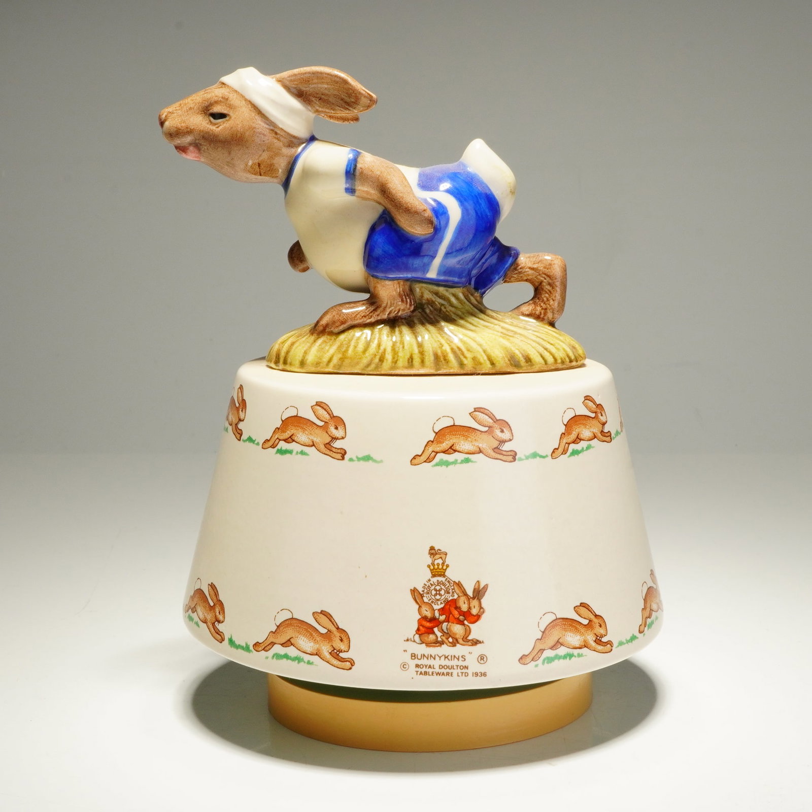 Royal Doulton Jogging Bunnykins Music Box DB37: This Royal Doulton Bunnykins music box figurine features the Jogging Bunnykins DB37 model, often associated with the tune King of the Road. The piece consists of a ceramic topper depicting a rabbit in