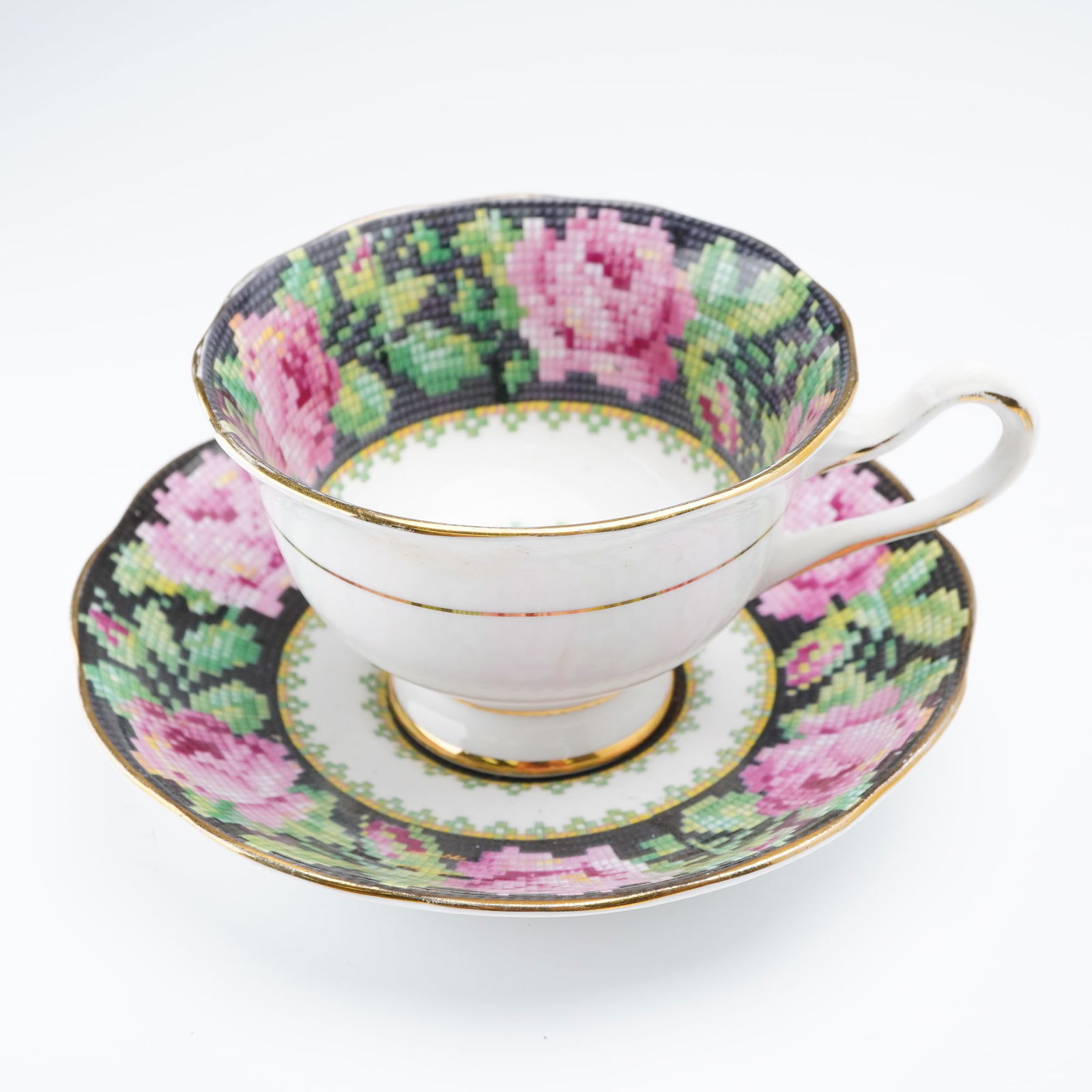 Royal Albert Porcelain Cup & Saucer Set, Needle Point: A charming cup and saucer set in the Needle Point pattern. The design features a cross-stitch-style motif of vibrant pink roses and green foliage set against a dark ground, bordered in fine gold gilt.