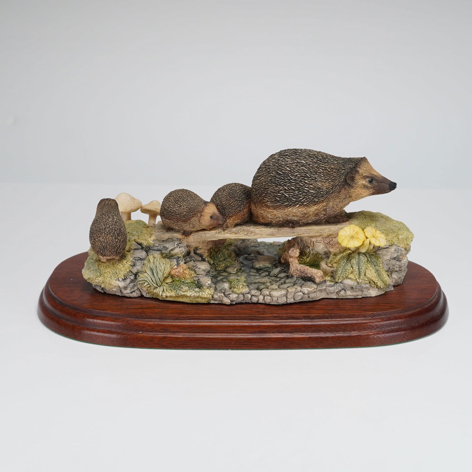 Border Fine Arts Hedgehogs Figurine: A detailed wildlife sculpture titled 'Hedgehogs' by Border Fine Arts. The composition features four hedgehogs (one adult and three young) resting on a log that spans a simulated stream and rocky bank,