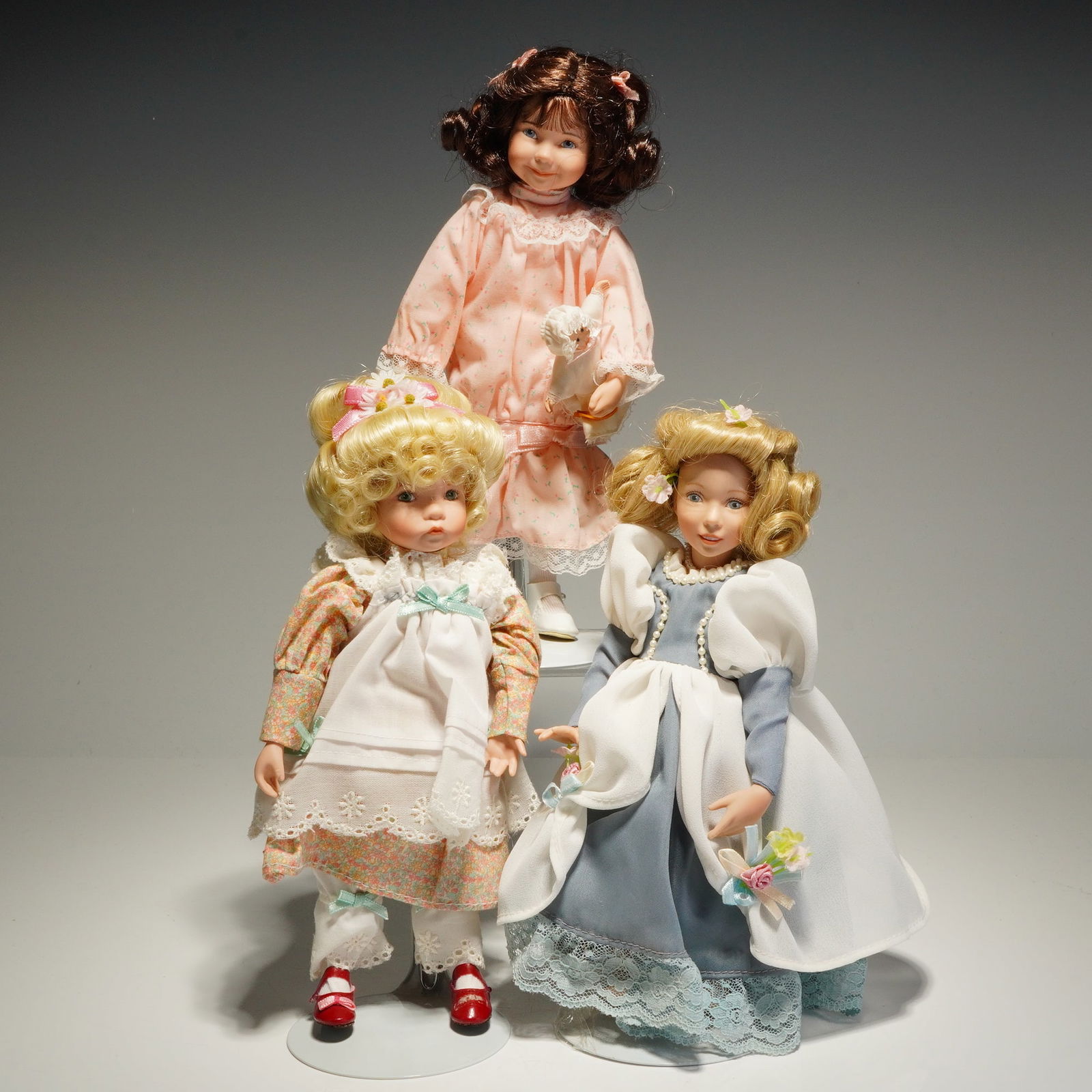 3pc Dianna Effner Porcelain Collectible Dolls: This group of dolls are constructed with ceramic heads, arms, and legs attached to soft bodies. The collection includes a dark-haired doll in a pink floral dress holding a miniature baby doll, a