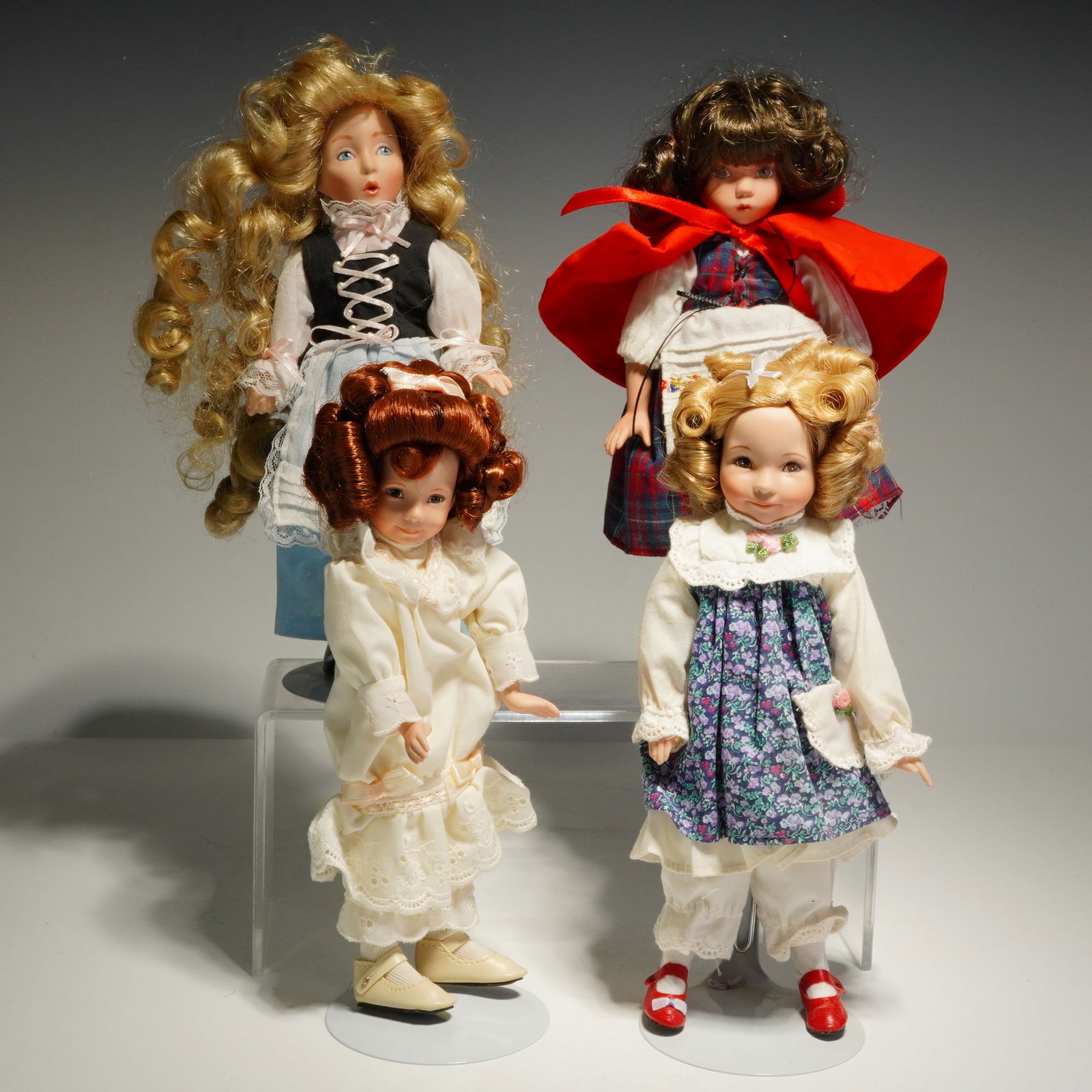 4pc Dianna Effner Porcelain Dolls: This collection of dolls showcases bisque porcelain heads, arms, and legs combined with soft bodies. Each doll is dressed in detailed fabric costumes, featuring styles such as a red hooded cape over a
