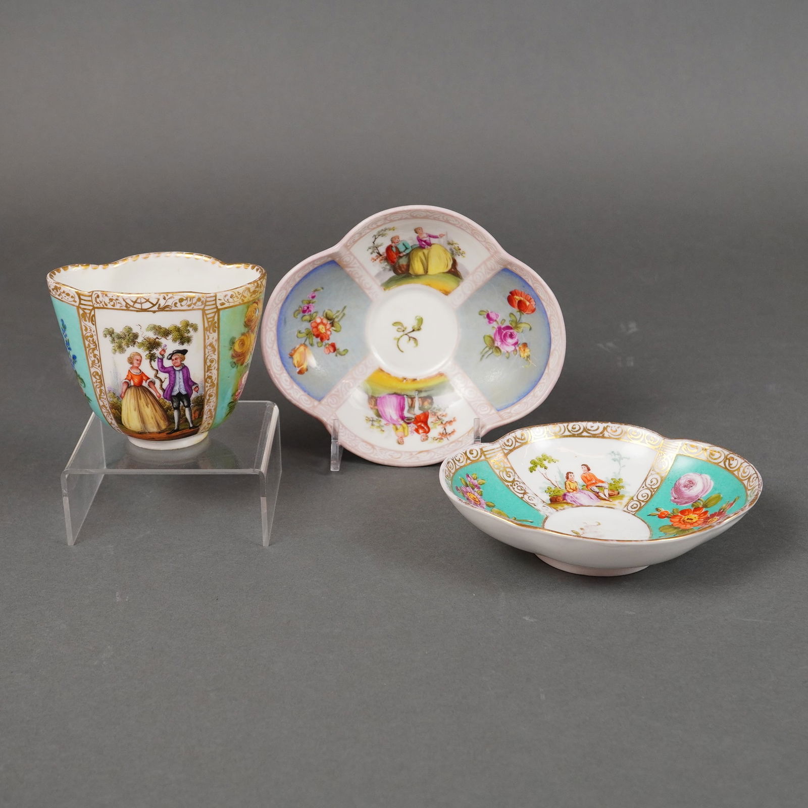 A Trio of Augustus Rex Style Porcelain Watteau-Decorated Items: A trio of European porcelain items executed in the Rococo style, comprising one handleless cup and two quatrefoil-shaped dishes or saucers. The decoration is polychrome, featuring hand-painted