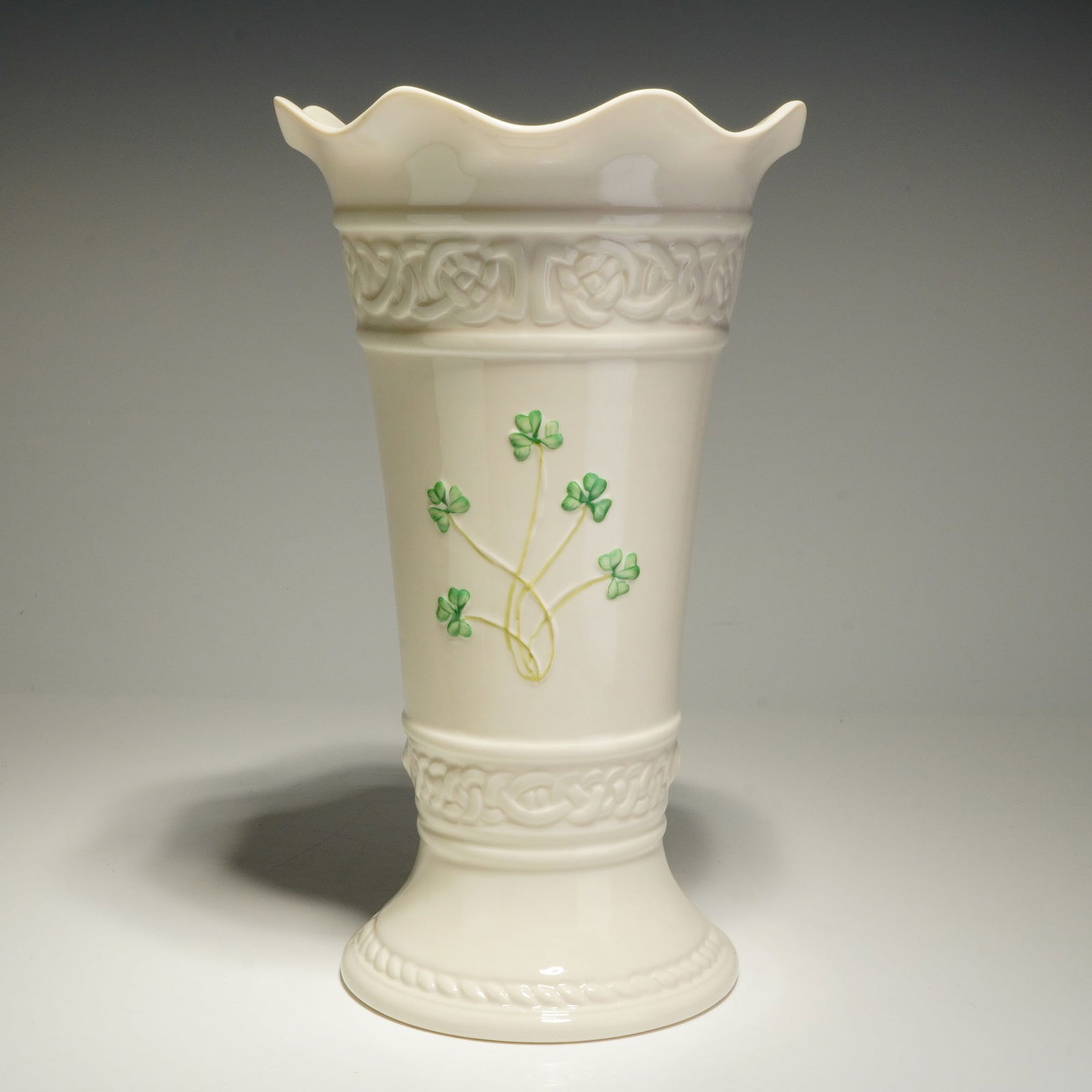 Belleek Classic Tara Shamrock Vase: This Belleek Parian china vase features a flared, cylindrical body with a scalloped rim and a pedestal base. The cream-colored vessel is decorated with embossed Celtic knotwork bands around the upper