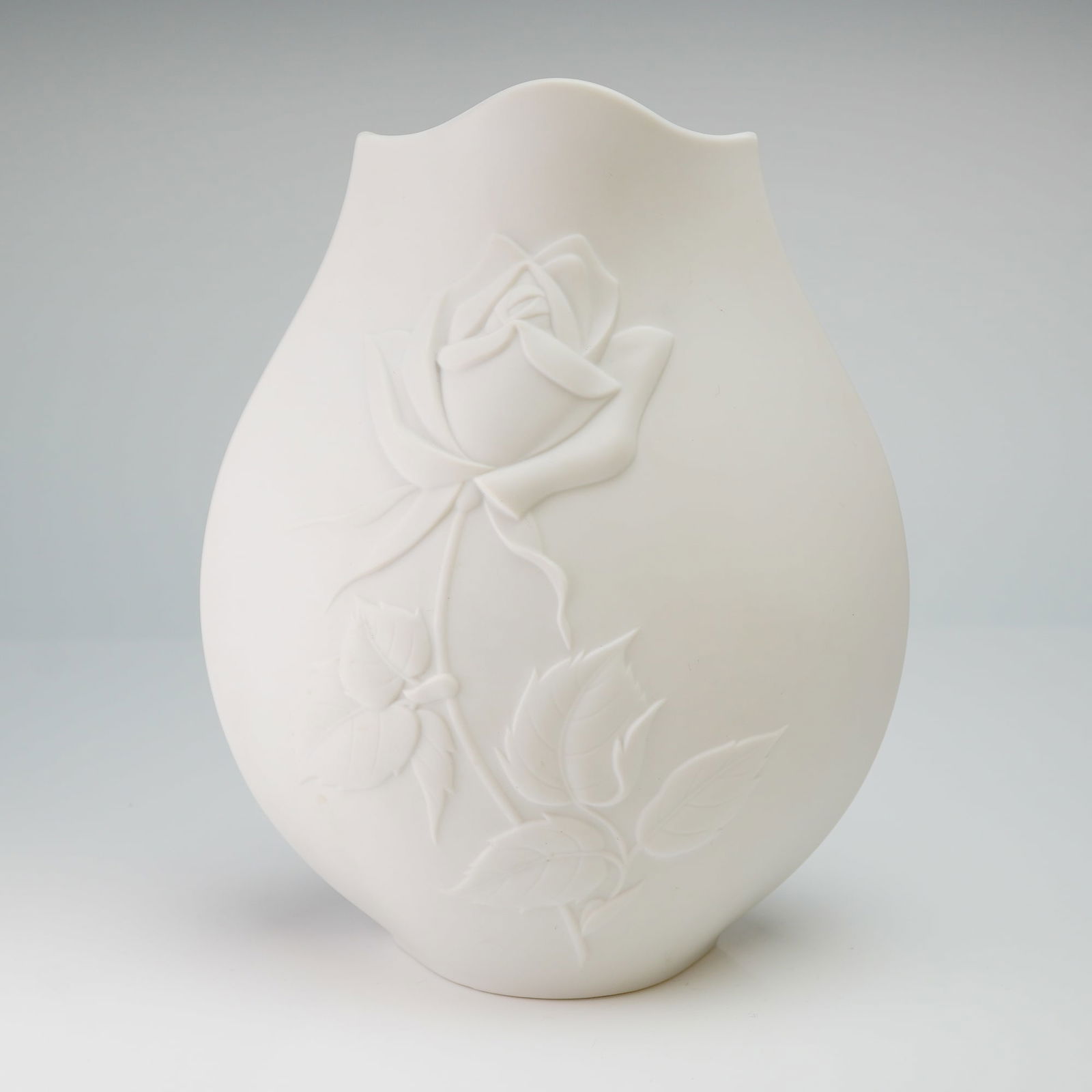 Noritake White Bisque Porcelain Rose Vase: The vessel features a bulbous, teardrop-shaped body terminating in a decorative scalloped rim. The obverse is adorned with a prominent bas-relief molded rose, complete with stem and foliage. The under