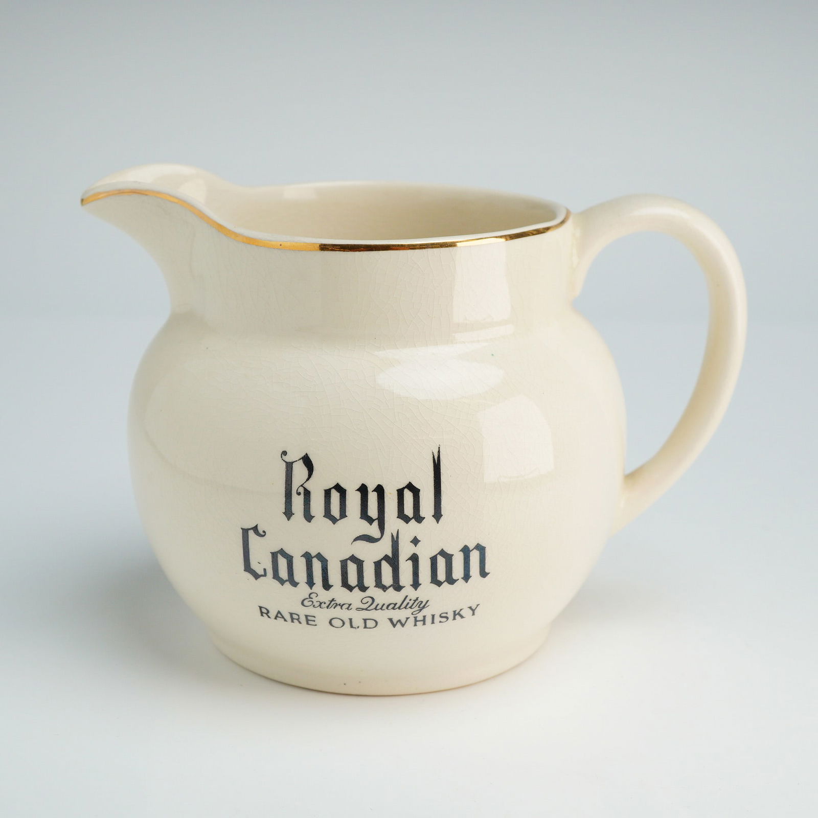 Wade Porcelain Pitcher for Royal Canadian Whisky: The advertising vessel features a globular body with a cream-colored glaze and gilt trim along the rim and handle. The front of the jug displays black transfer-printed text reading Royal Canadian