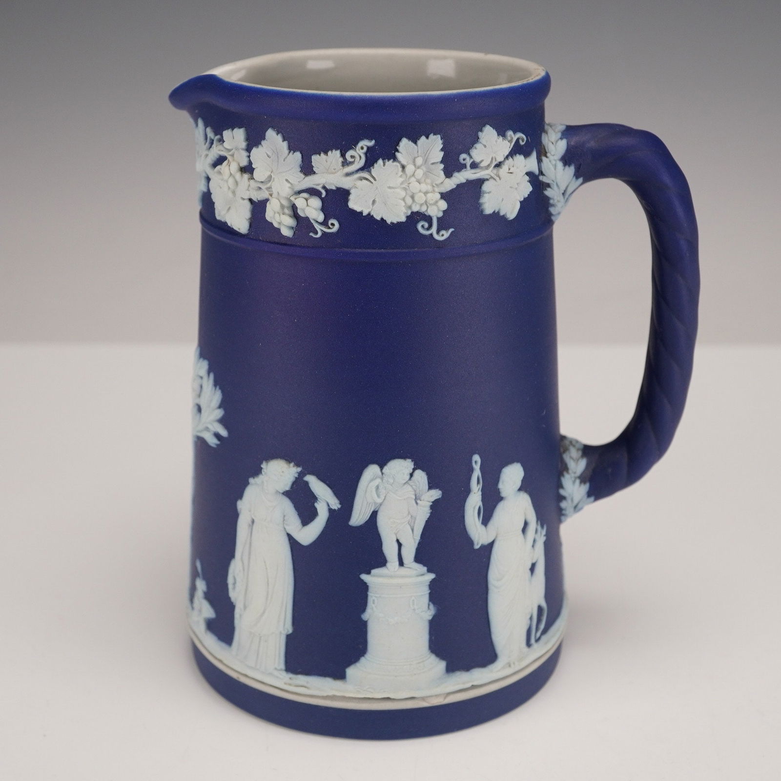 A Wedgwood Neo-Classical Dark Blue Jasperware Jug: A Wedgwood Neo-Classical Dark Blue Jasperware Jug, featuring a matte deep blue exterior with intricate white bas-relief decoration. The design includes a continuous frieze of grapevines and clusters a