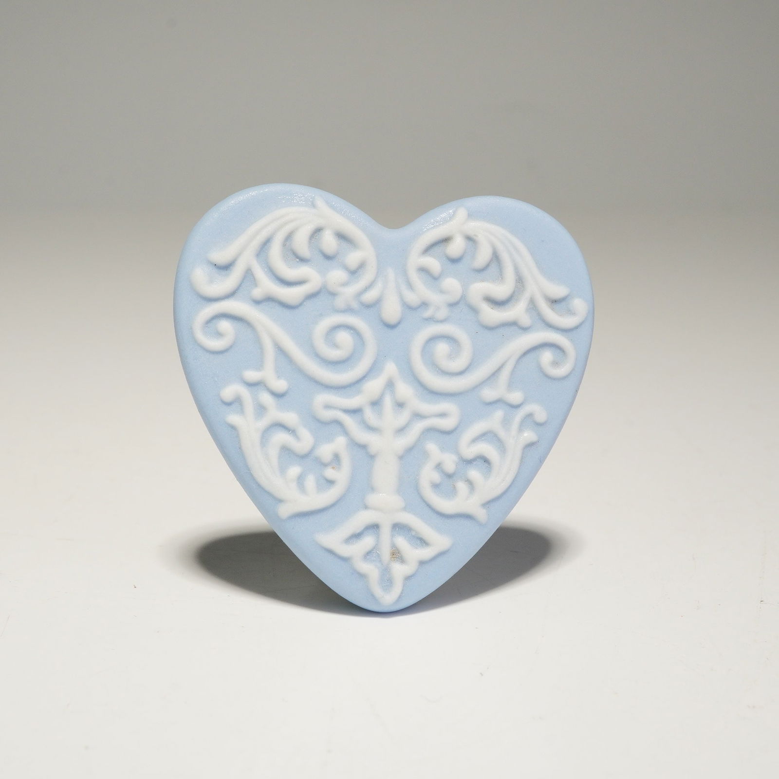 Wedgwood Blue Jasperware Heart Brooch Pin: A Wedgwood blue jasperware heart-shaped brooch featuring a light blue ground with white bas-relief filigree and foliate patterns. The reverse side bears the printed mark WEDGWOOD with a registered tra