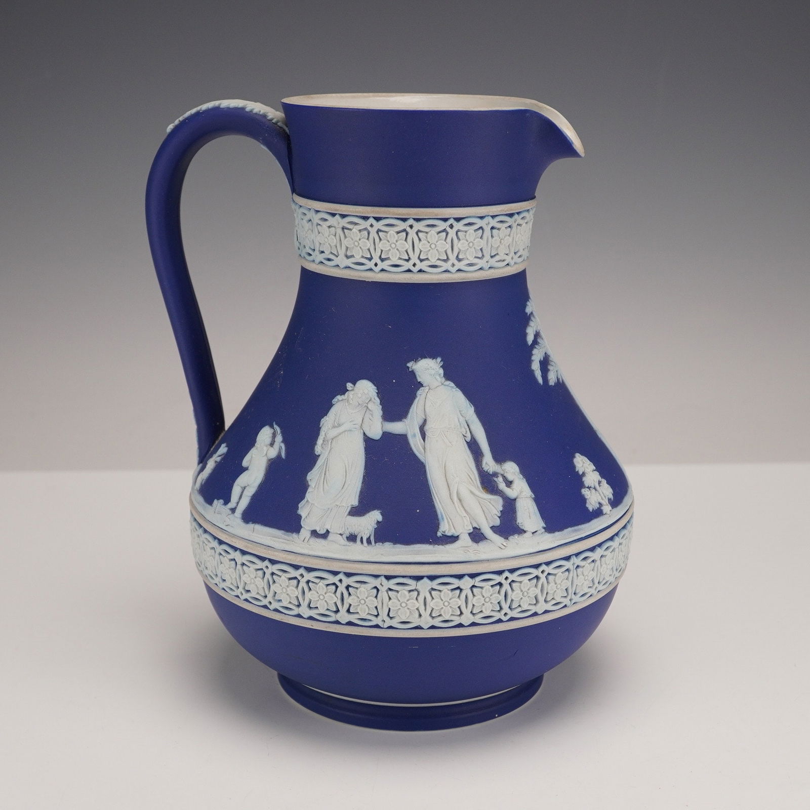 Wedgwood Dark Blue Jasperware Pitcher: A Wedgwood Dark Blue Jasperware Pitcher with a deep blue matte, unglazed finish characteristic of jasperware. Its ovoid body is richly decorated with white bas-relief classical figures, depicting drap