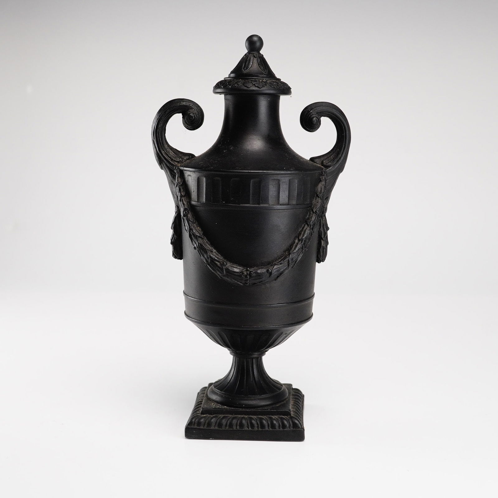 Wedgwood Black Basalt Covered Urn: A small urn-shaped vase with lid, crafted in Black Basalt. The body is adorned with relief-molded drapery garlands and fluted detailing along the shoulder and foot. Two scrolled handles frame the vess