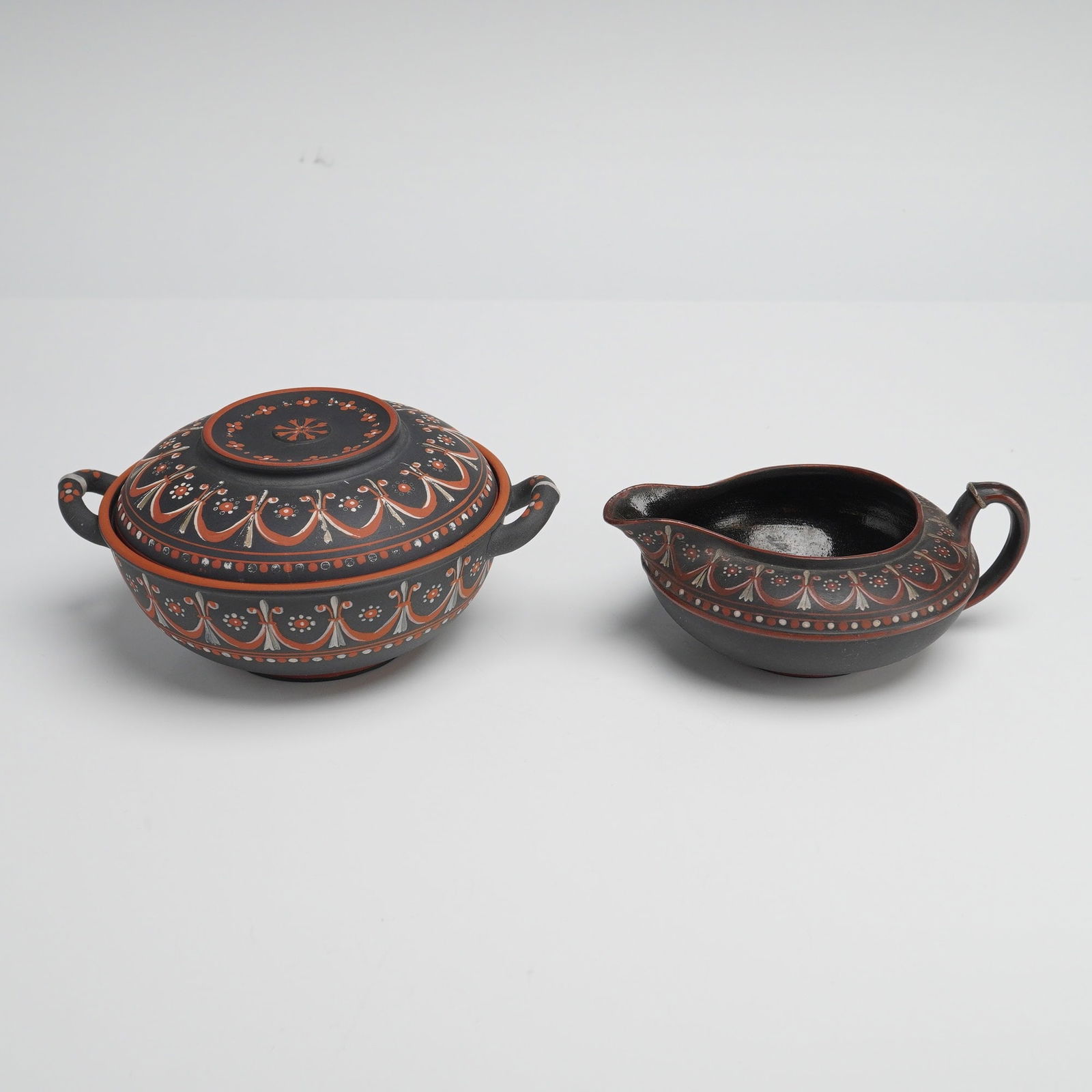 Two Wedgwood Encaustic Decorated Black Basalt Items: Two Wedgwood encaustic-decorated black basalt items, England, late 18th century, comprising a lidded two-handled sugar bowl and a single-handled cream pitcher. The pieces are crafted from matte black