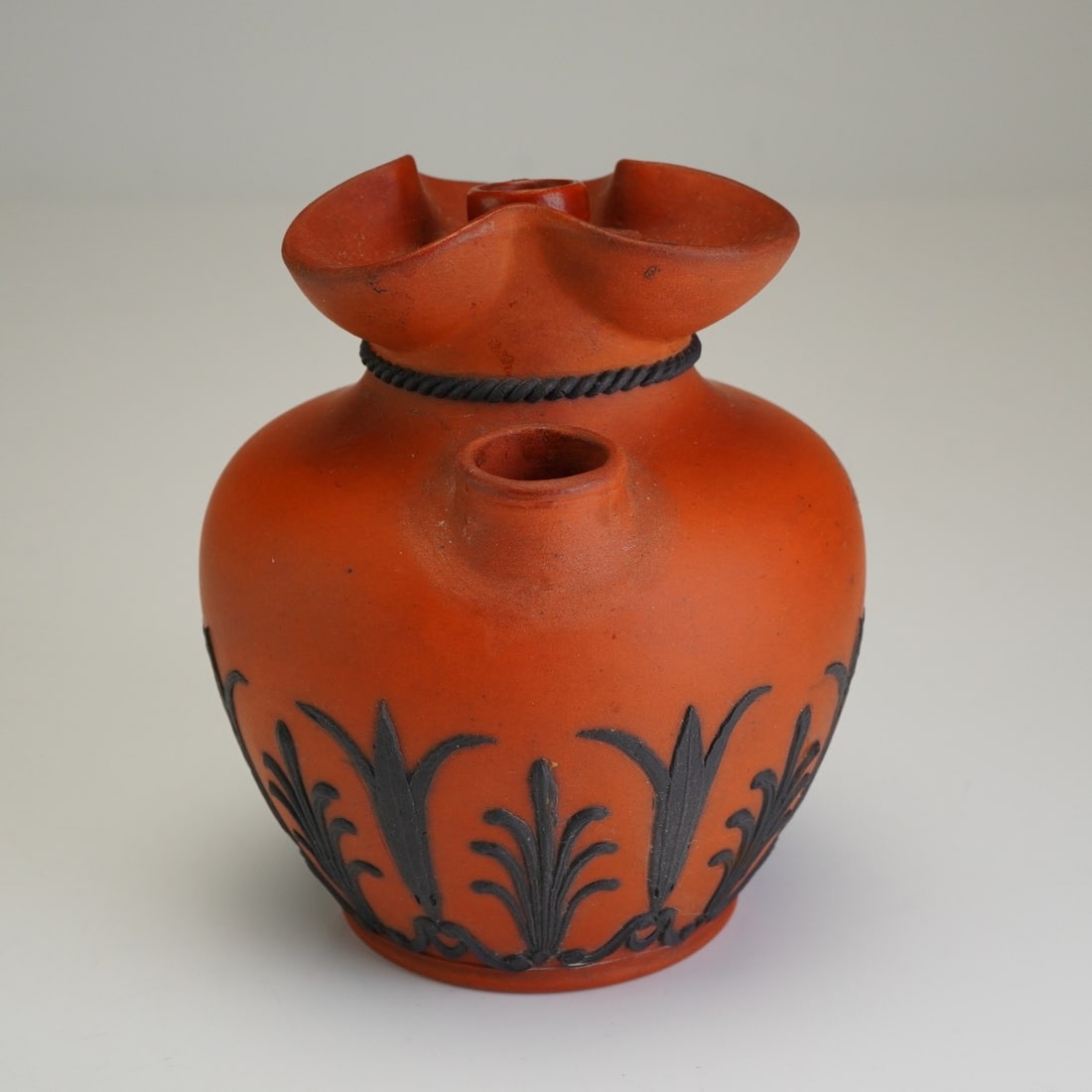 Wedgwood Rosso Antico and Black Basalt Pyryphorous Vase: The vessel features a bulbous body with a trilobed, ruffled rim and a single circular spout on one shoulder, with two elongated oval apertures on the opposite side. The decoration consists of black ba
