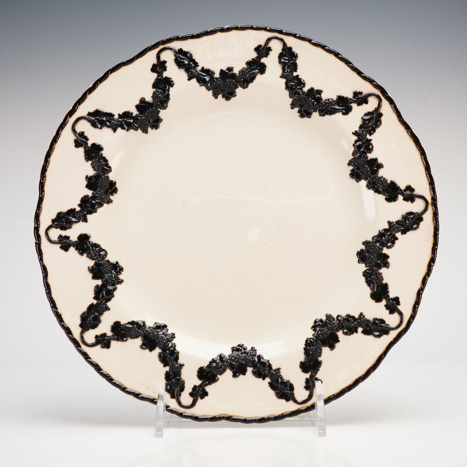 Wedgwood Creamware Plate with Black Basalt Bas-Relief Decoration: A Wedgwood creamware plate featuring a glossy cream-colored glaze and a scalloped rim. The interior is decorated with a continuous black border and a raised, sprigged grapevine garland in black basalt