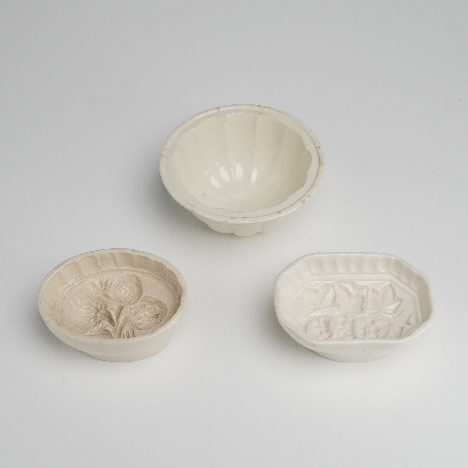 Three Wedgwood Staffordshire Creamware Jelly Molds: A trio of Wedgwood Staffordshire creamware jelly molds. The collection consists of three distinct ceramic forms: a circular fluted mold, an oval mold with a relief design depicting a cluster of strawb